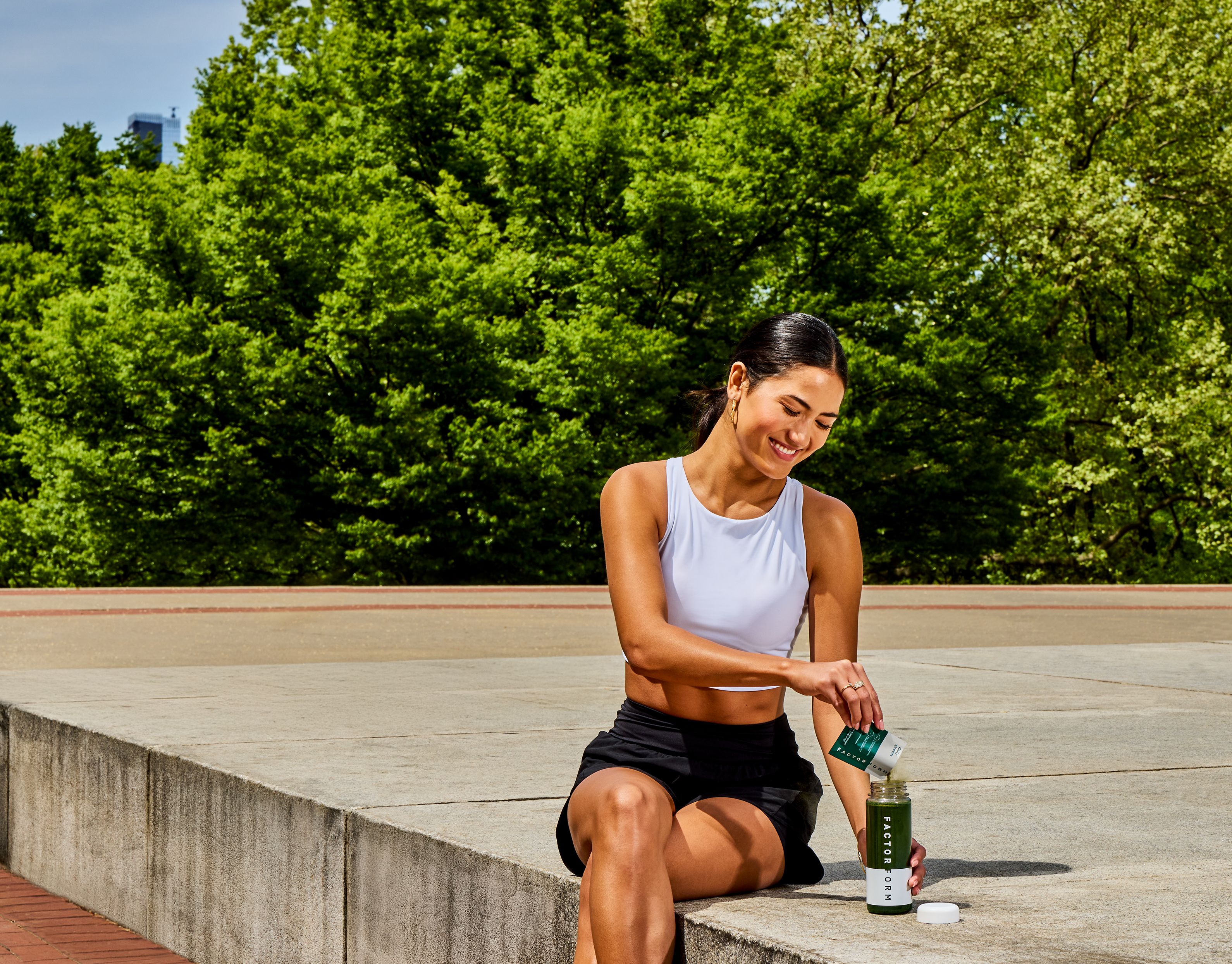 Smiling woman in athletic wear pouring powder into a Factor 88 shaker bottle outdoors.