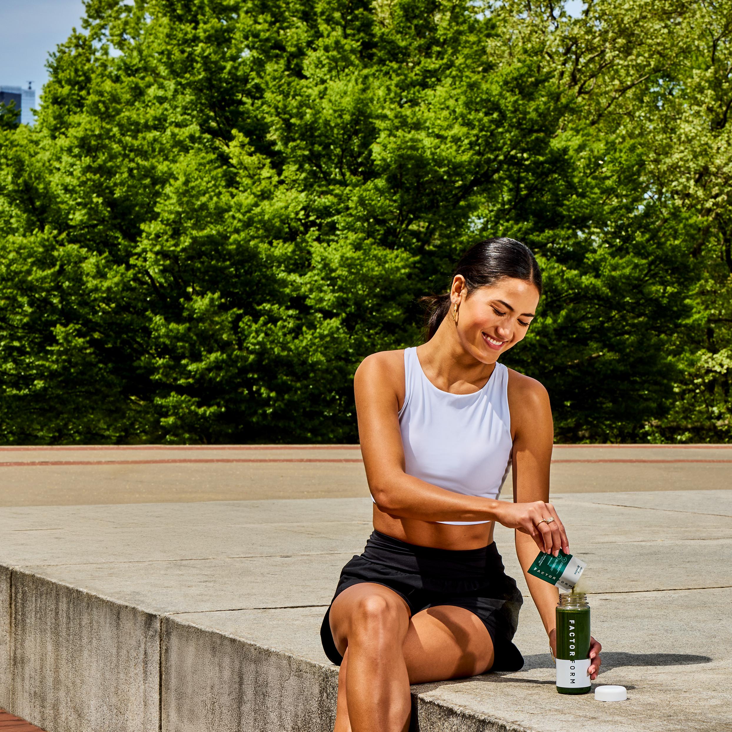 Smiling woman in athletic wear pouring powder into a Factor 88 shaker bottle outdoors.
