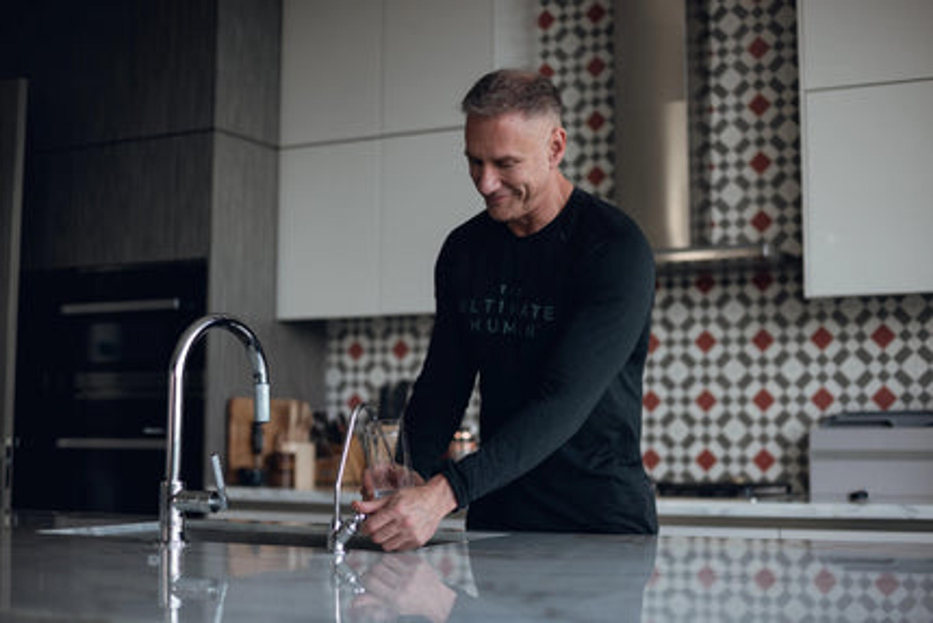 A man in an "ULTIMATE HUMAN" shirt fills a glass with water from a tap in a modern kitchen.