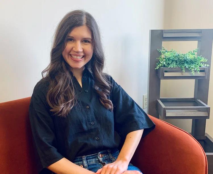 A smiling woman in a black shirt and jeans sits on an orange chair, with a wooden plant stand behind her.