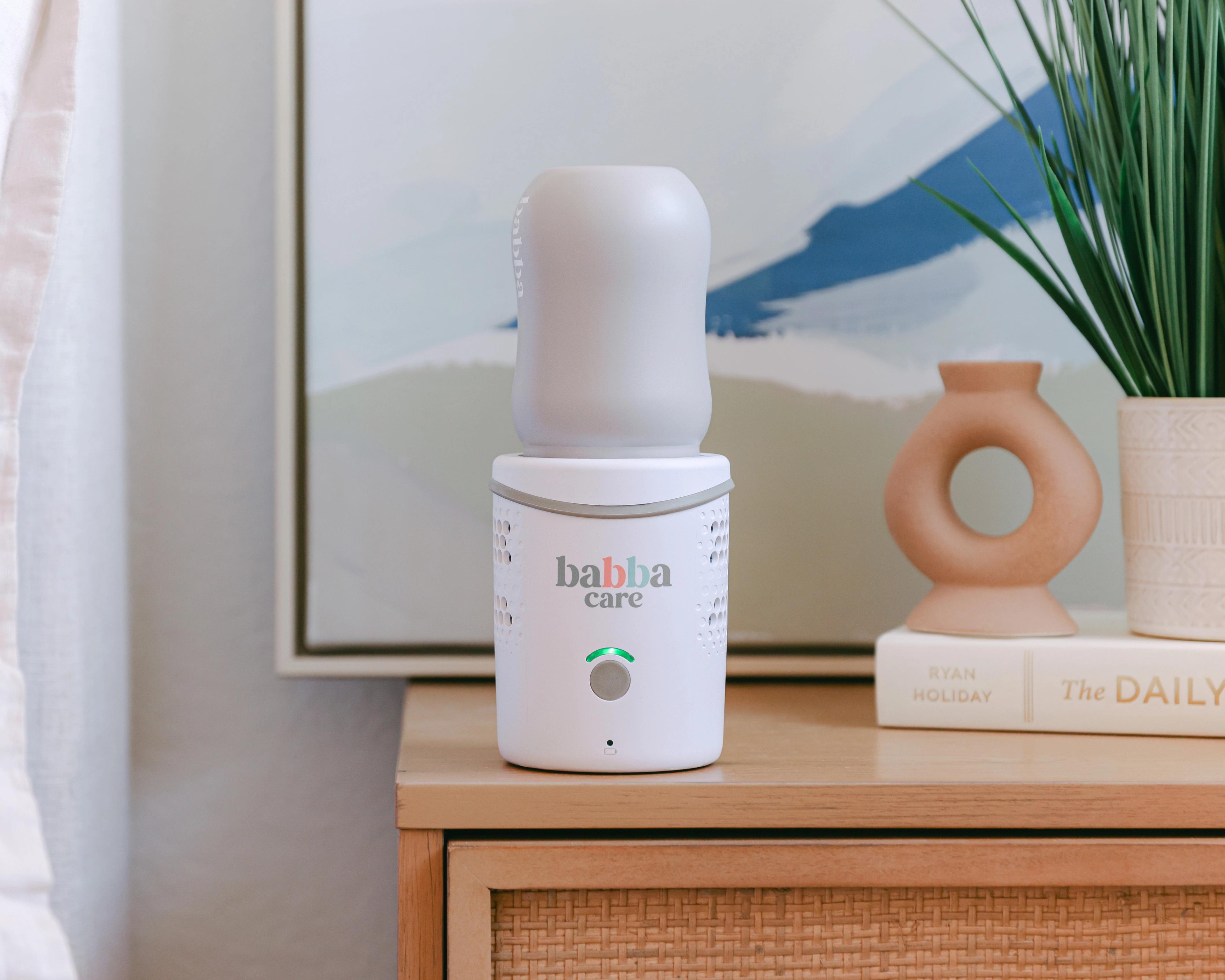 A white Babba Care baby formula maker sits on a wooden nightstand with decorative items in the background.