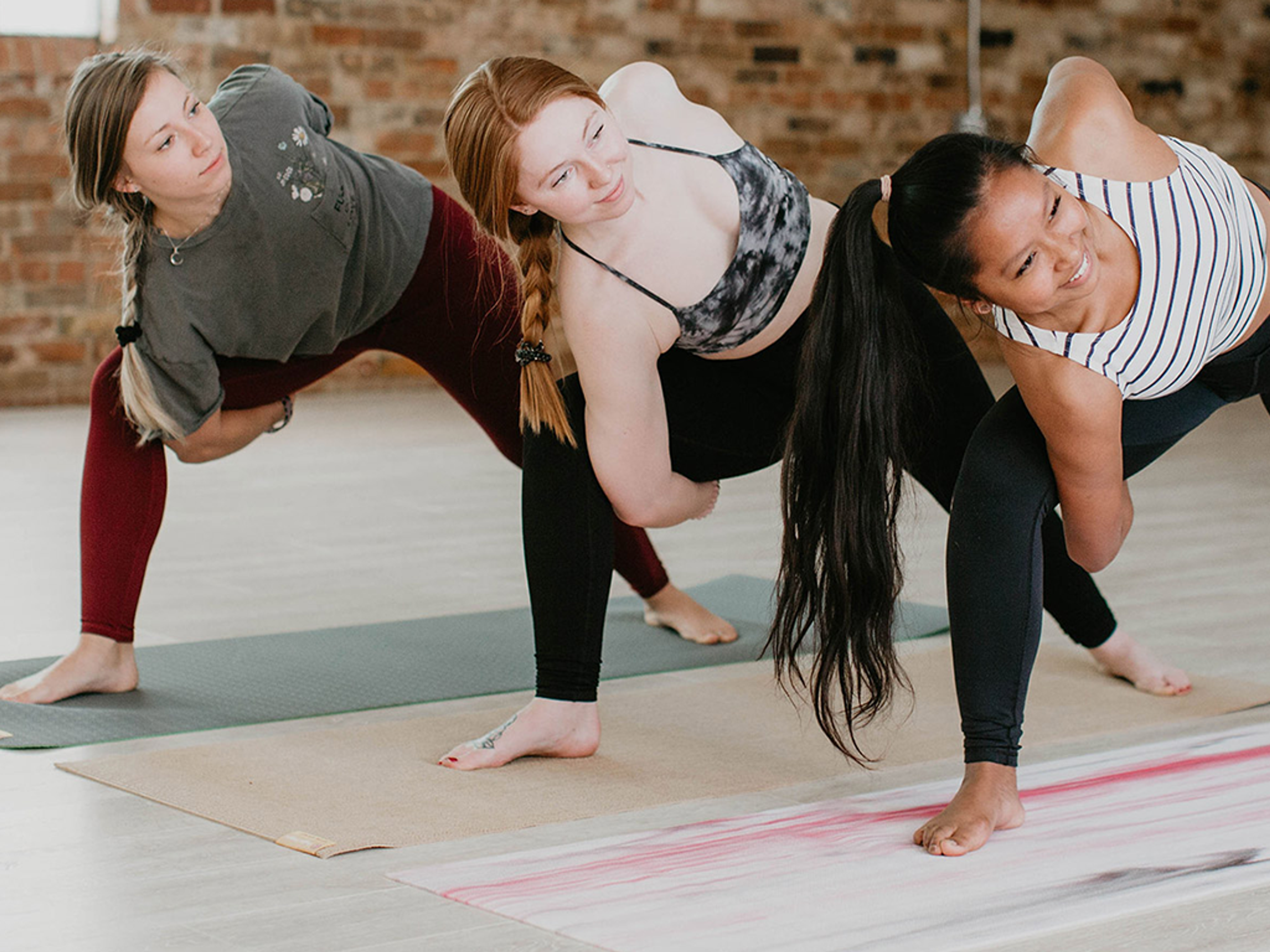 Three diverse women perform a revolved side angle yoga pose on mats.
