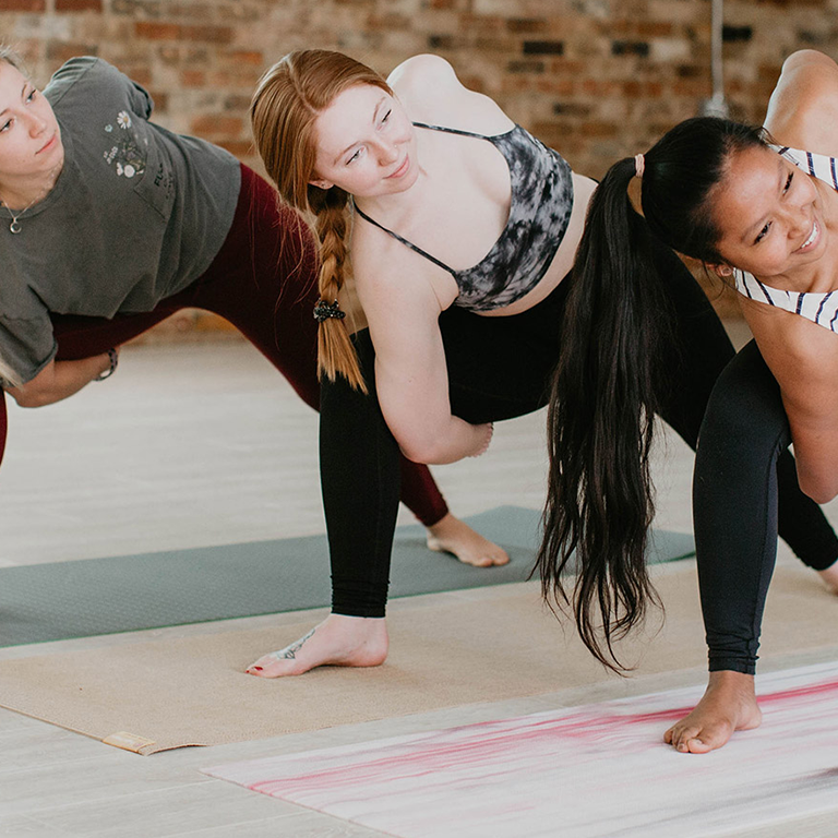 Three diverse women perform a revolved side angle yoga pose on mats.