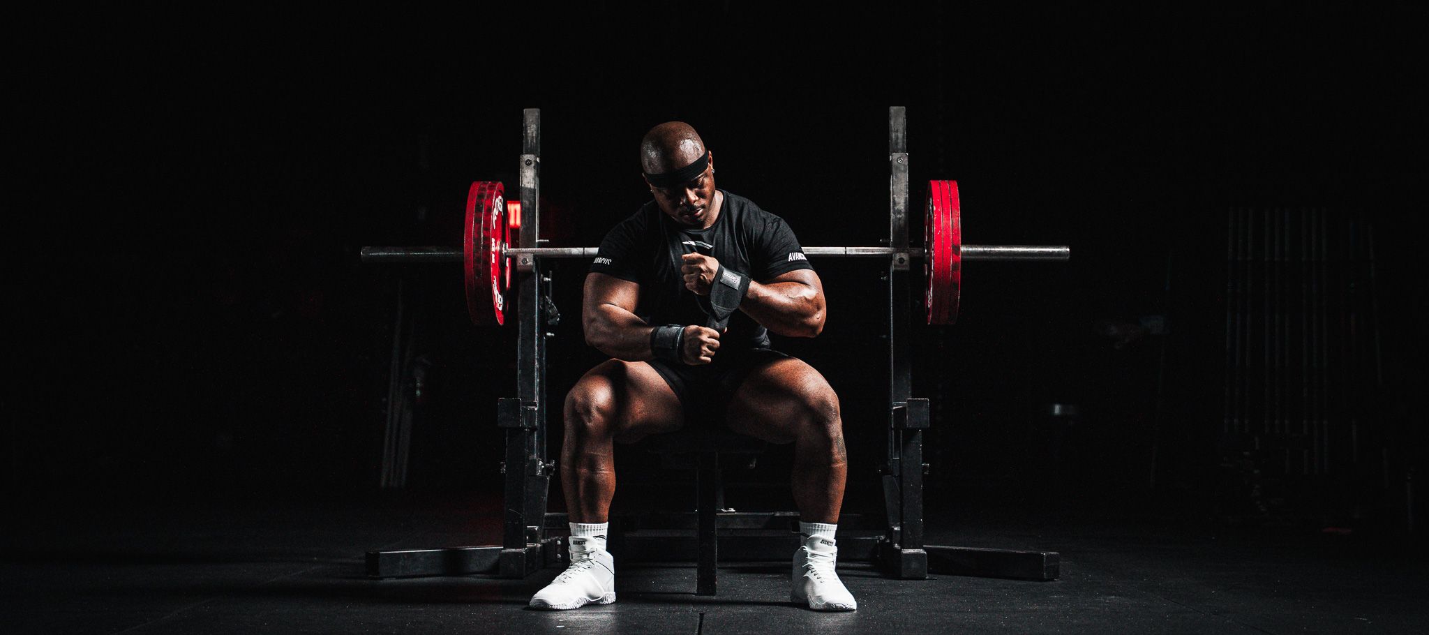 Muscular man adjusting wrist wraps while sitting in front of a barbell.