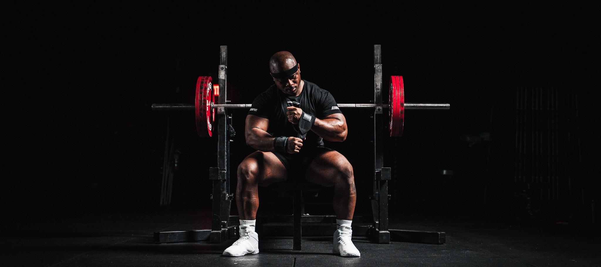 Muscular man adjusting wrist wraps while sitting in front of a barbell.