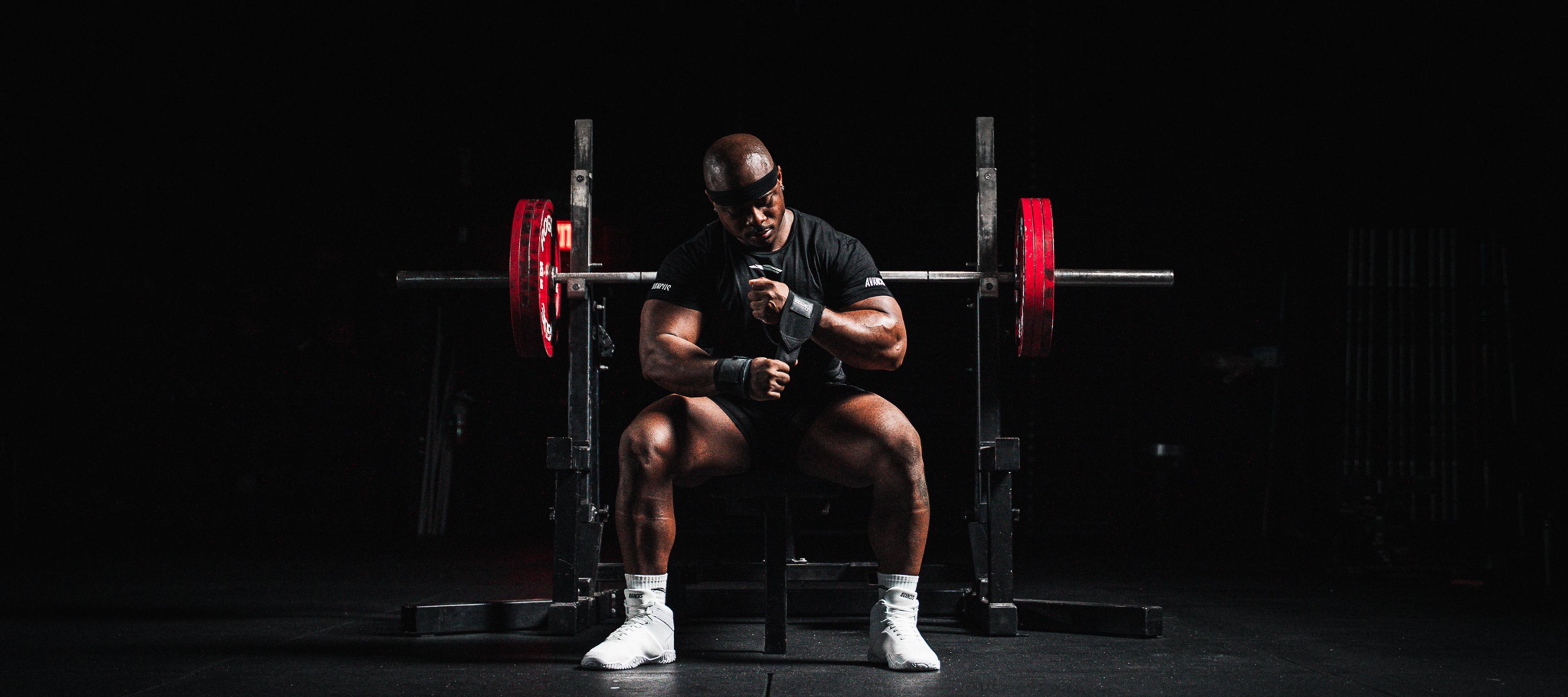 Muscular man adjusting wrist wraps while sitting in front of a barbell.