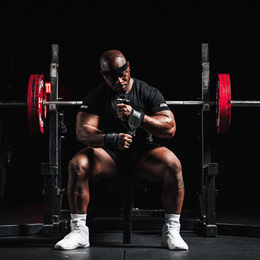 Muscular man adjusting wrist wraps while sitting in front of a barbell.