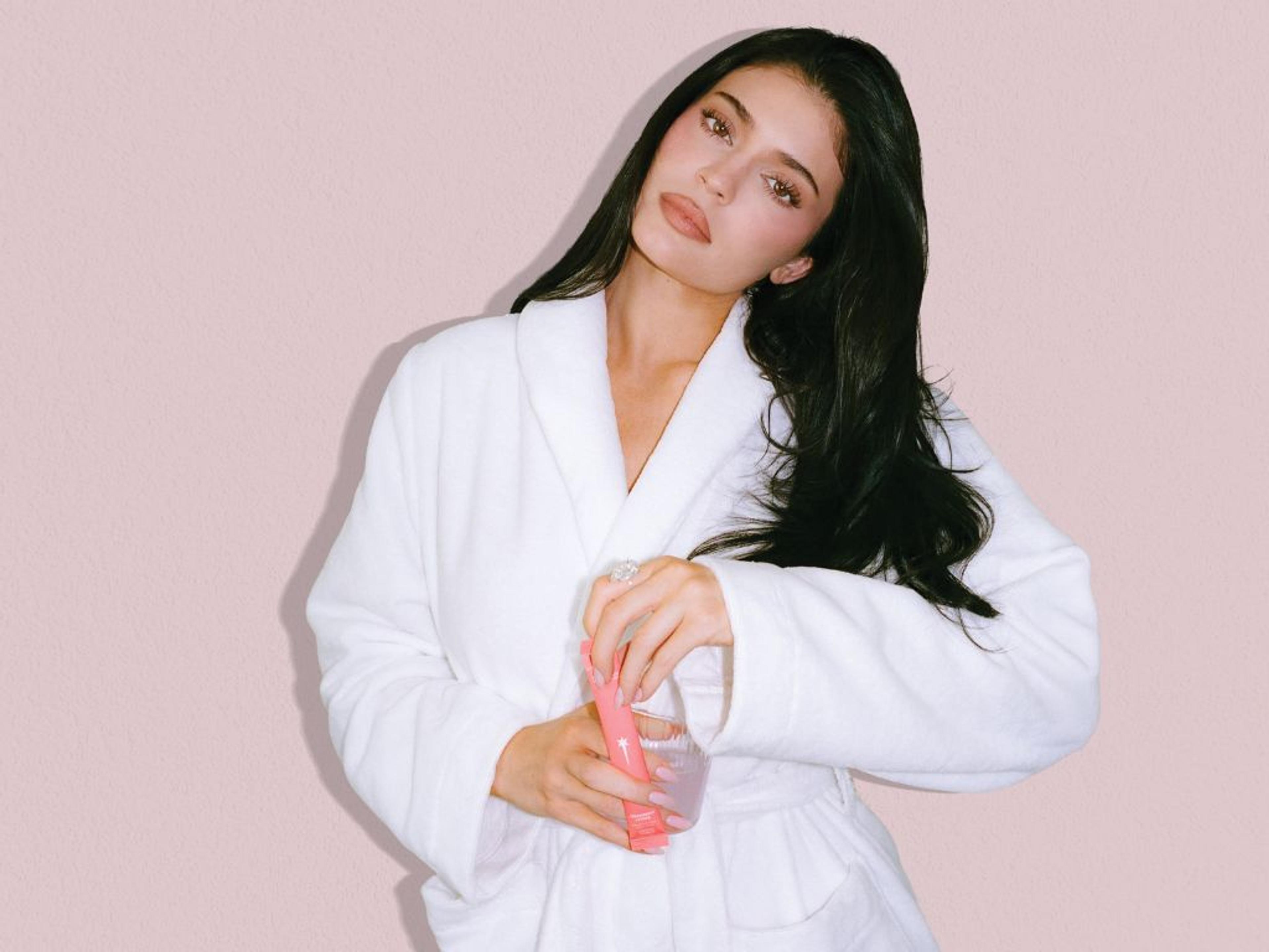 Kylie Jenner in a white robe, holding a pink tube and a glass with pink liquid.