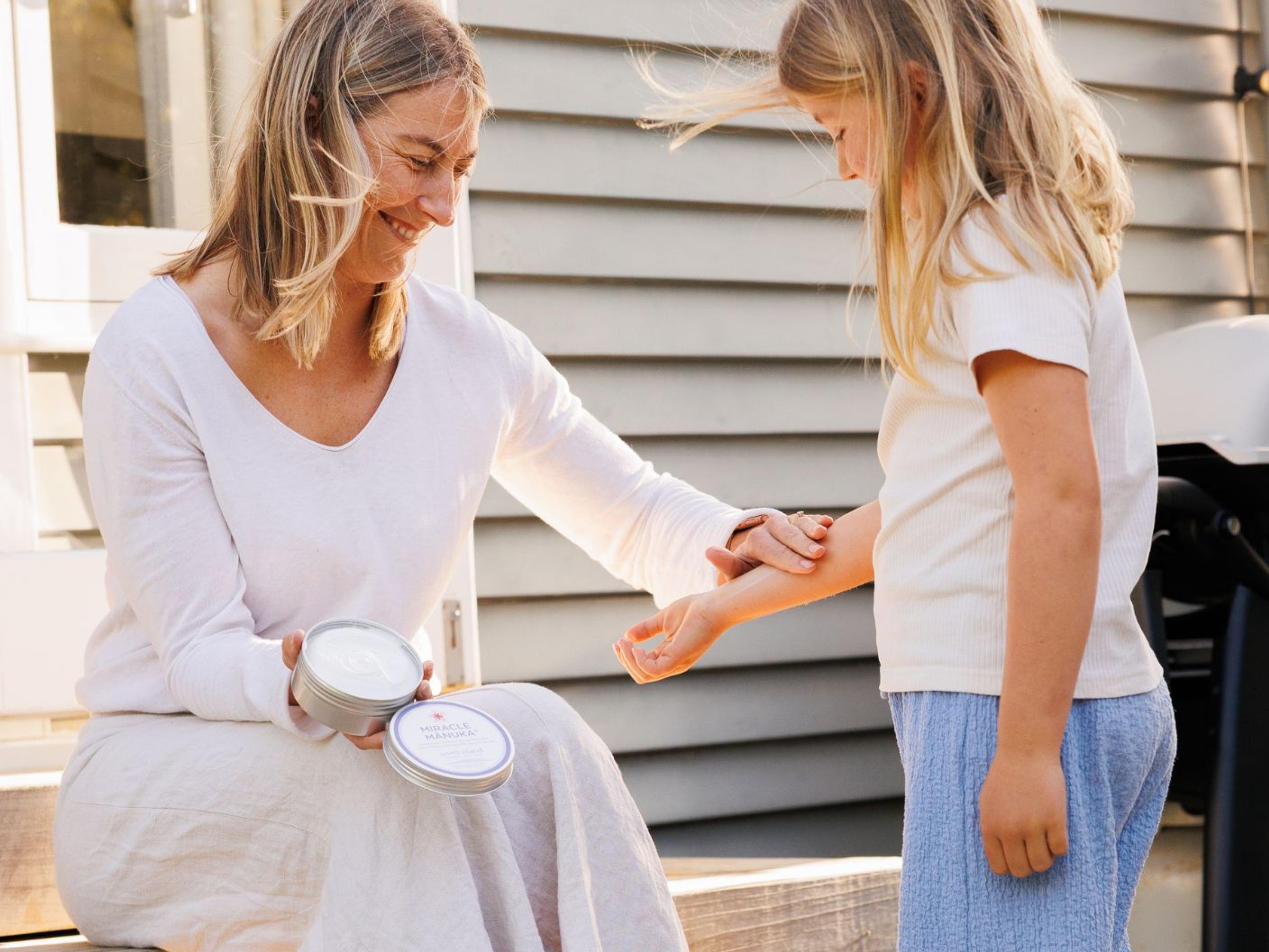 A smiling woman applies cream from a tin to a child's arm outdoors.