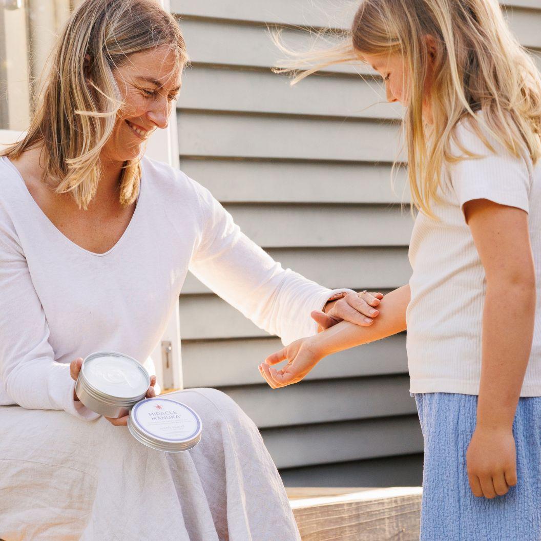 A smiling woman applies cream from a tin to a child's arm outdoors.
