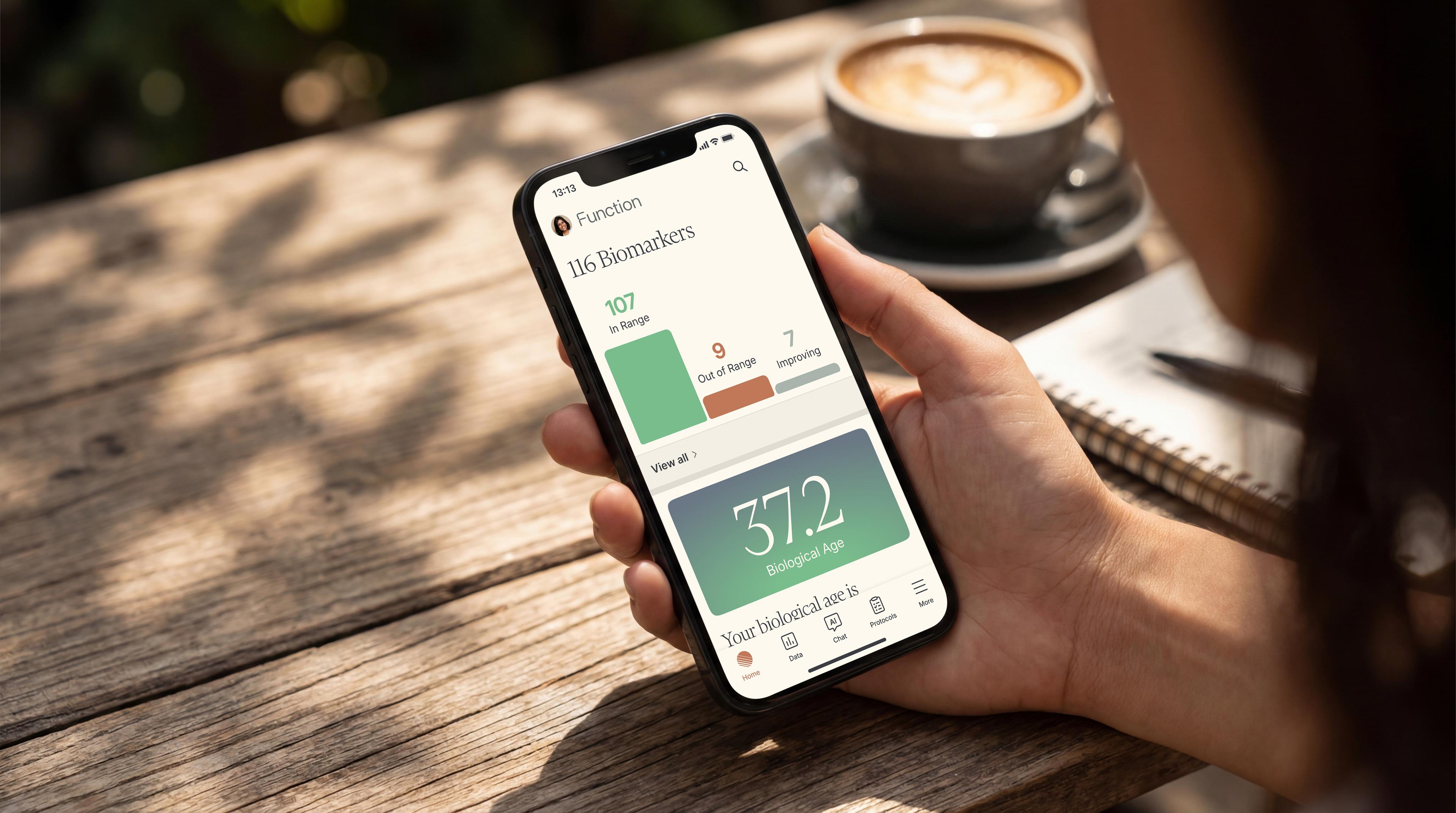 Hand holding a phone showing the Function app with 116 biomarkers tracked and a biological age of 37.2