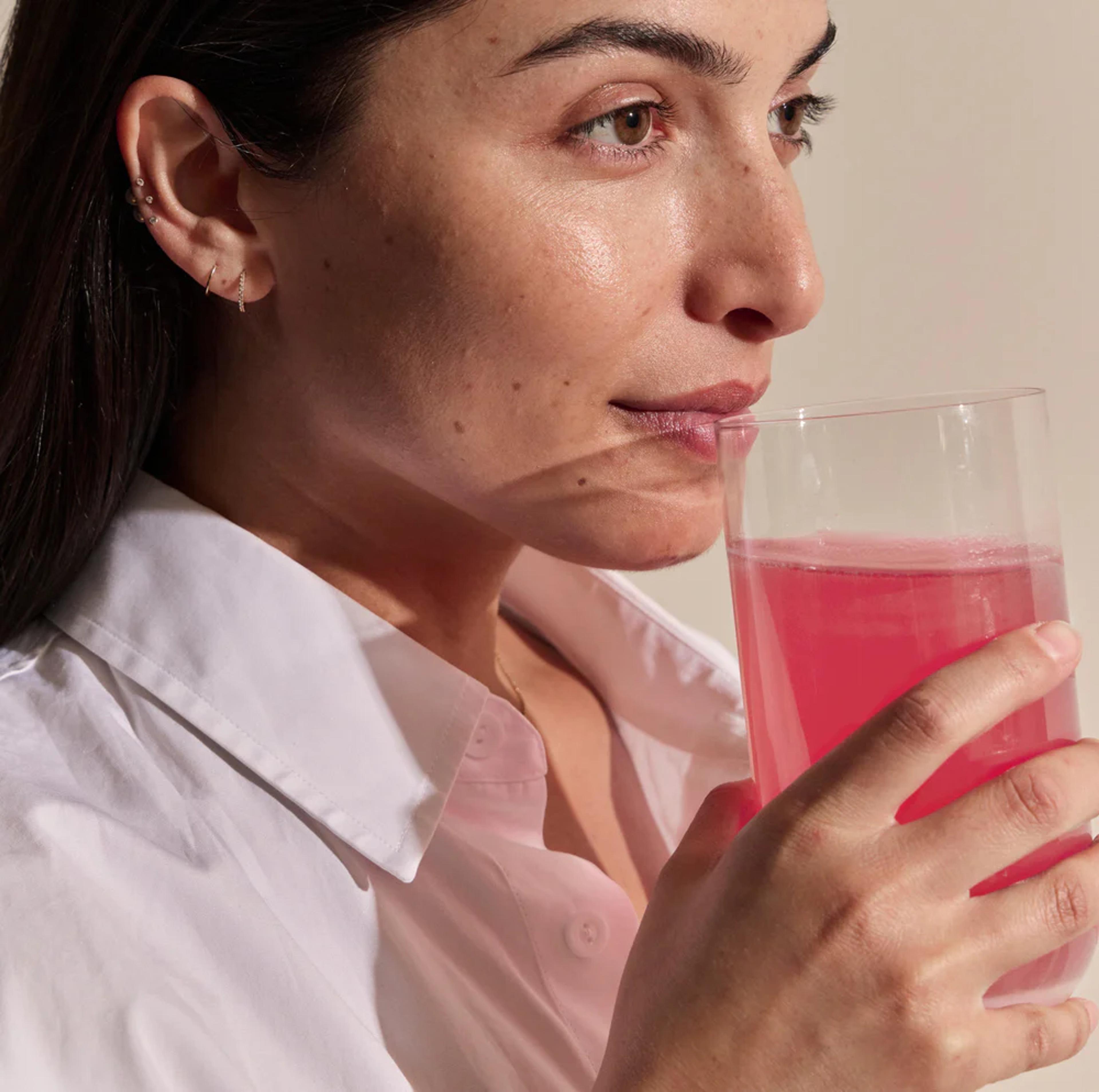 A woman in a white shirt drinks a pink beverage from a clear glass.