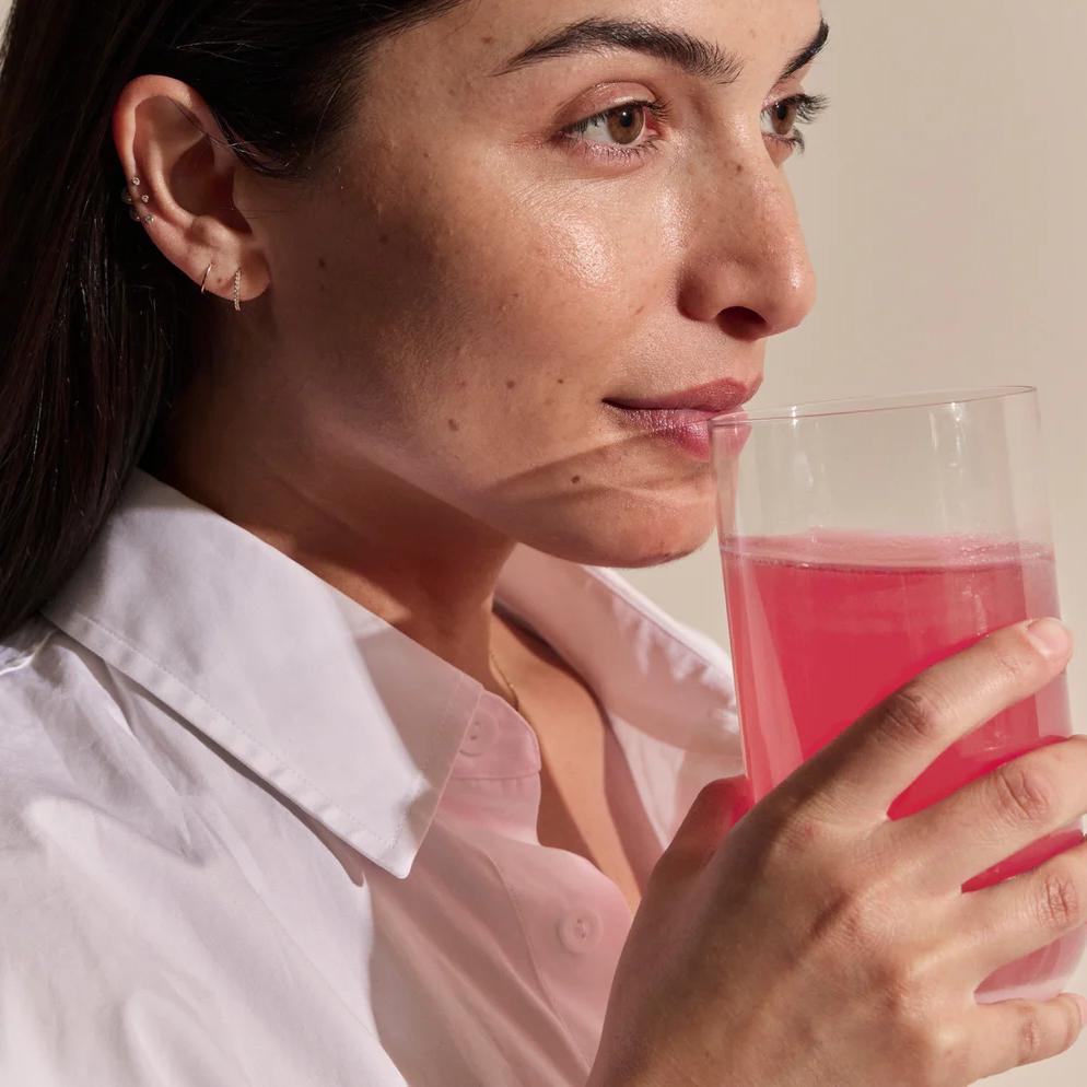 A woman in a white shirt drinks a pink beverage from a clear glass.