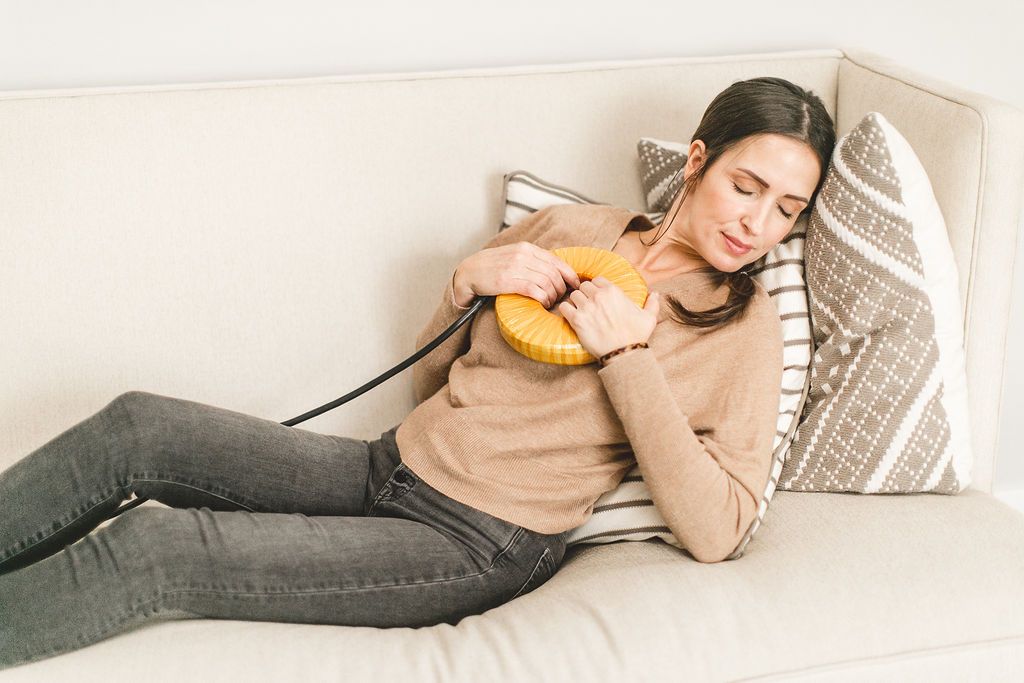 A relaxed woman lies on a sofa with eyes closed, holding a yellow, donut-shaped device with a cord to her chest.
