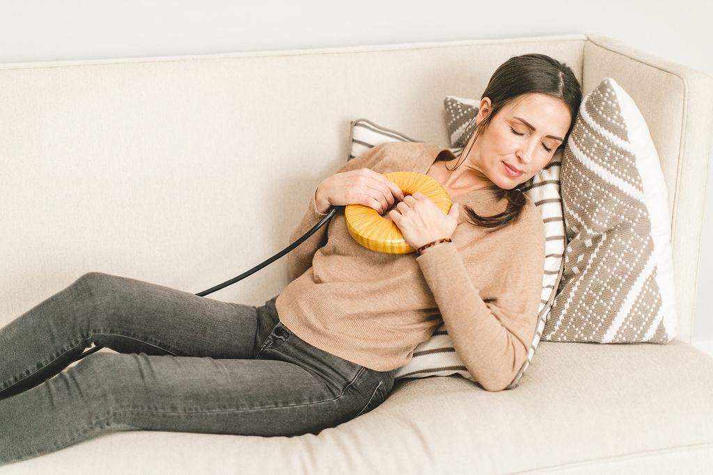 A relaxed woman lies on a sofa with eyes closed, holding a yellow, donut-shaped device with a cord to her chest.