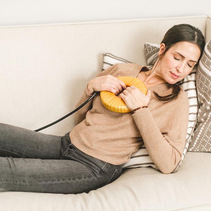 A relaxed woman lies on a sofa with eyes closed, holding a yellow, donut-shaped device with a cord to her chest.