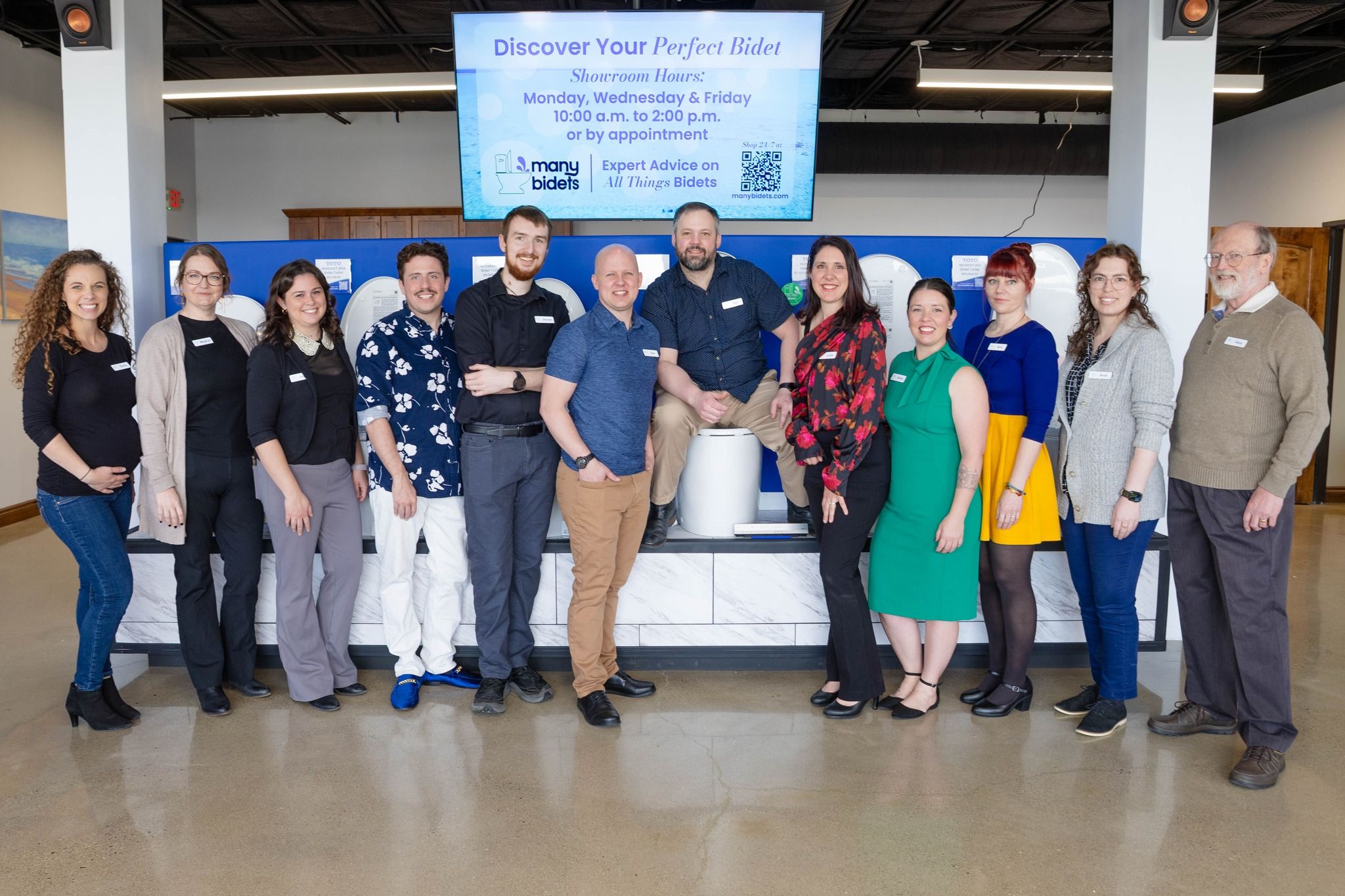 A diverse team of 12 people poses smiling in a bidet showroom, with one person seated on a bidet display. A screen above advertises "Discover Your Perfect Bidet."