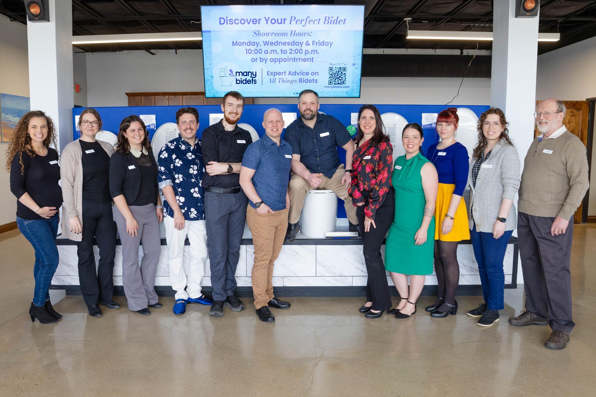 A diverse team of 12 people poses smiling in a bidet showroom, with one person seated on a bidet display. A screen above advertises "Discover Your Perfect Bidet."