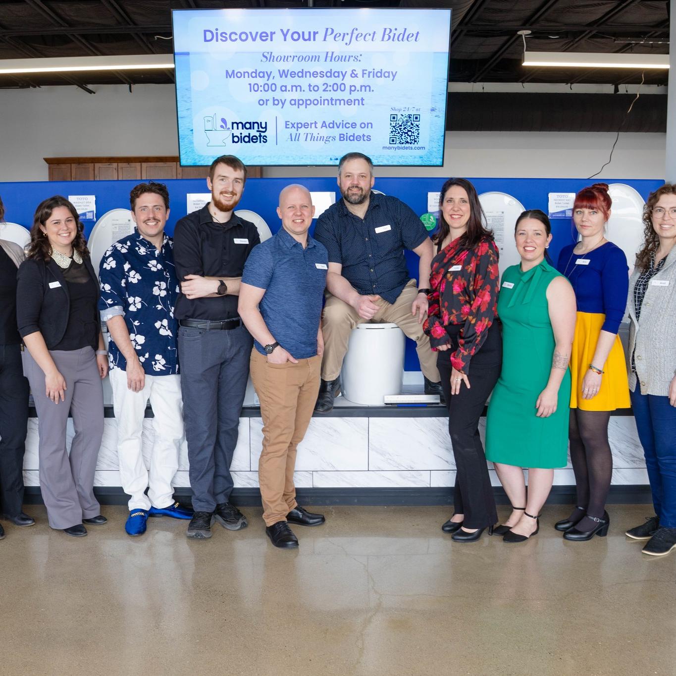 A diverse team of 12 people poses smiling in a bidet showroom, with one person seated on a bidet display. A screen above advertises "Discover Your Perfect Bidet."