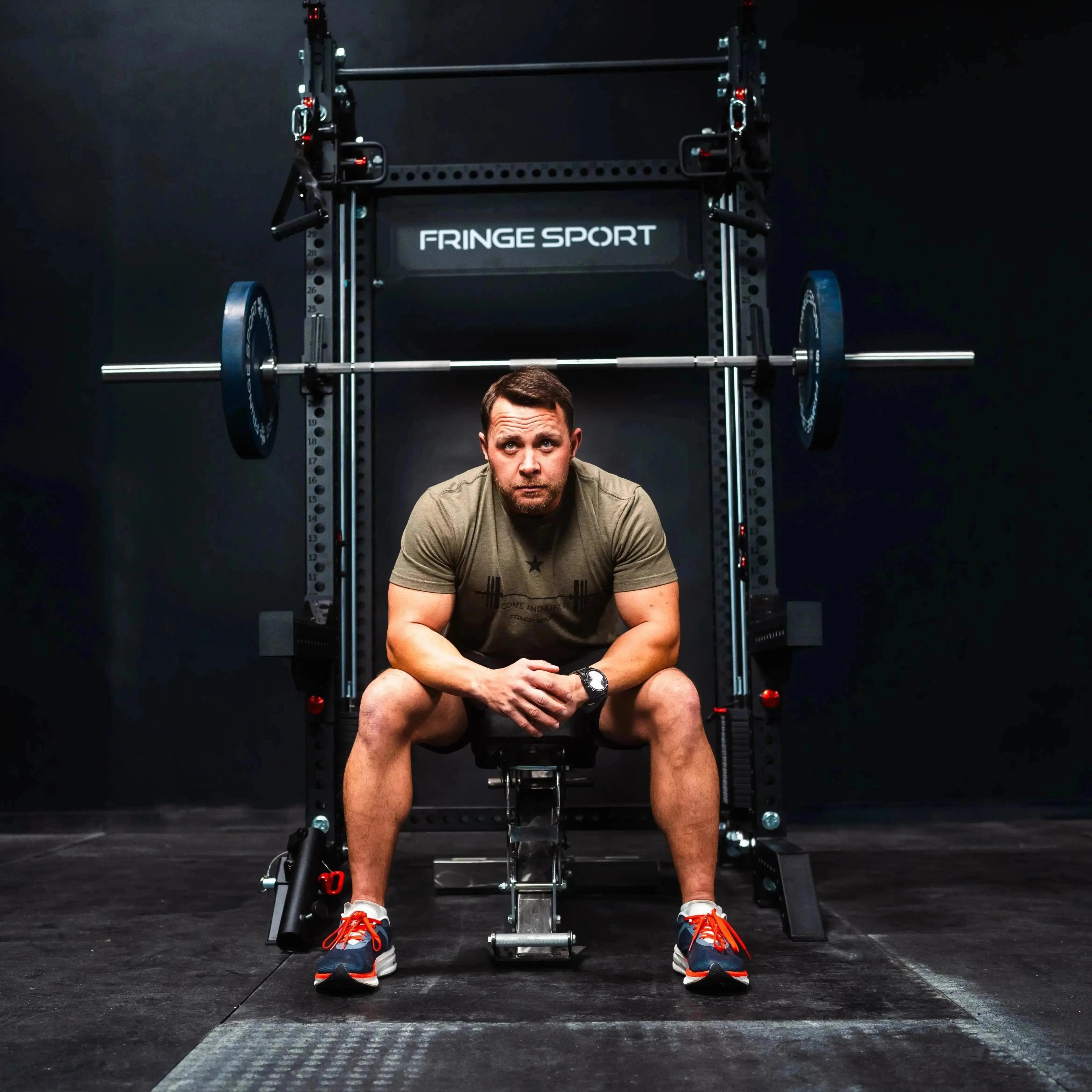 Man sitting on a bench in front of a Fringe Sport squat rack with a barbell.
