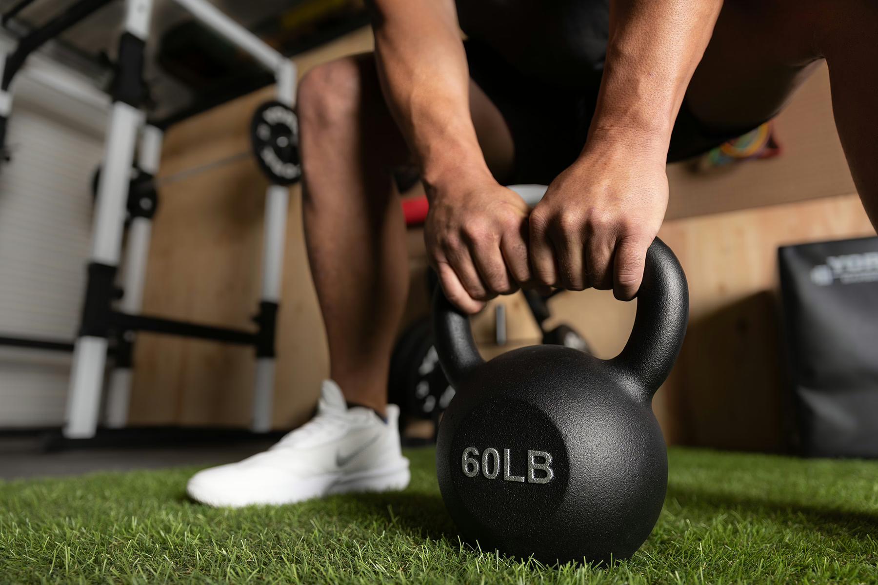 Person gripping a 60 lb black kettlebell on artificial turf.