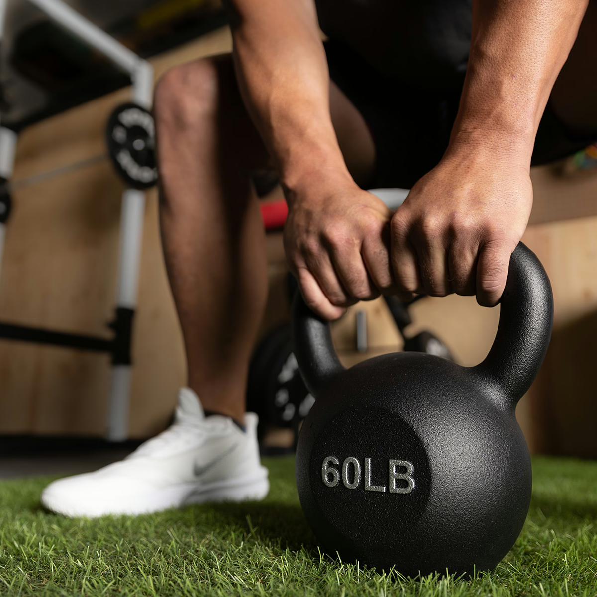 Person gripping a 60 lb black kettlebell on artificial turf.