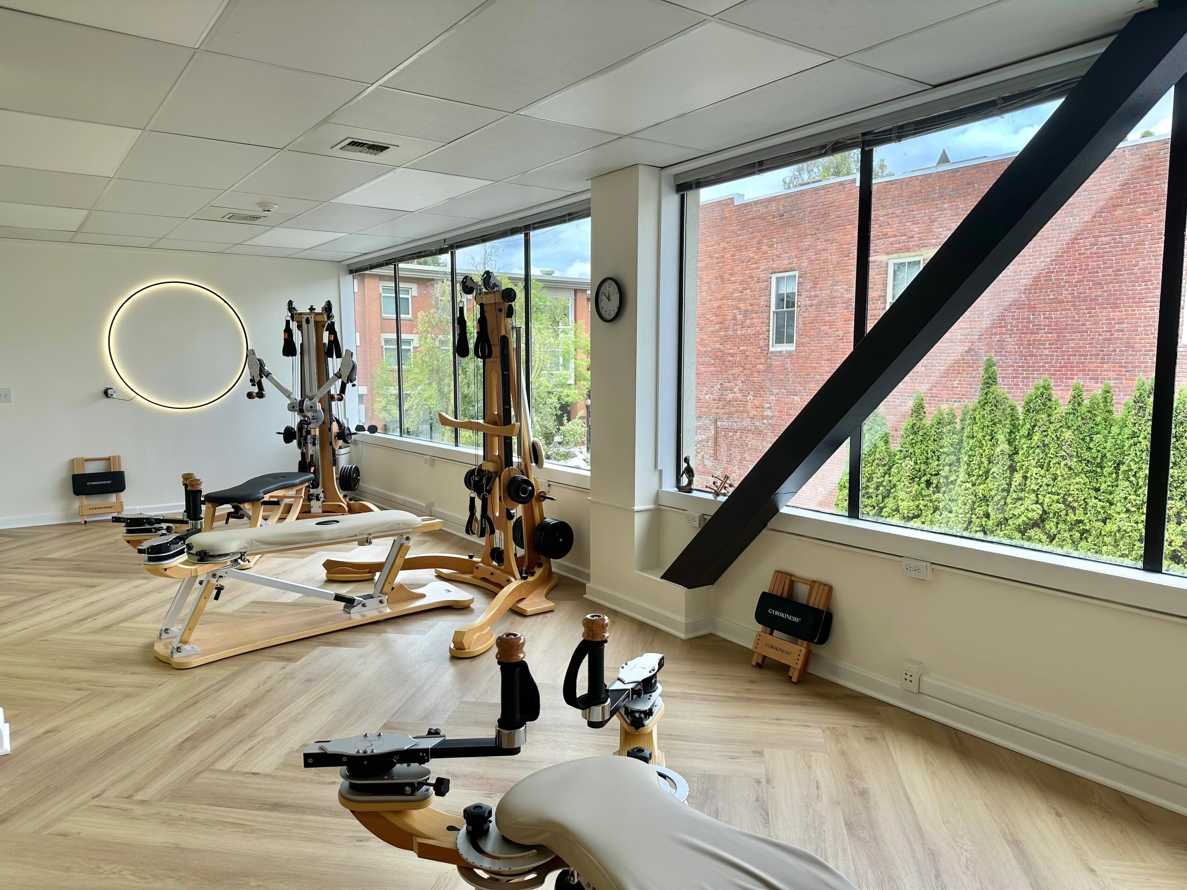 A bright room with light wood floors, several wooden exercise machines, a circular LED light, and large windows overlooking trees and a brick building.