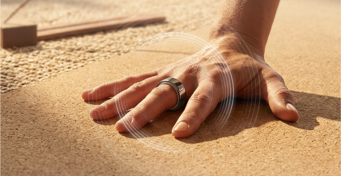 A hand wearing a silver smart ring on the index finger rests on a textured brown surface, with white signal waves radiating from the ring.