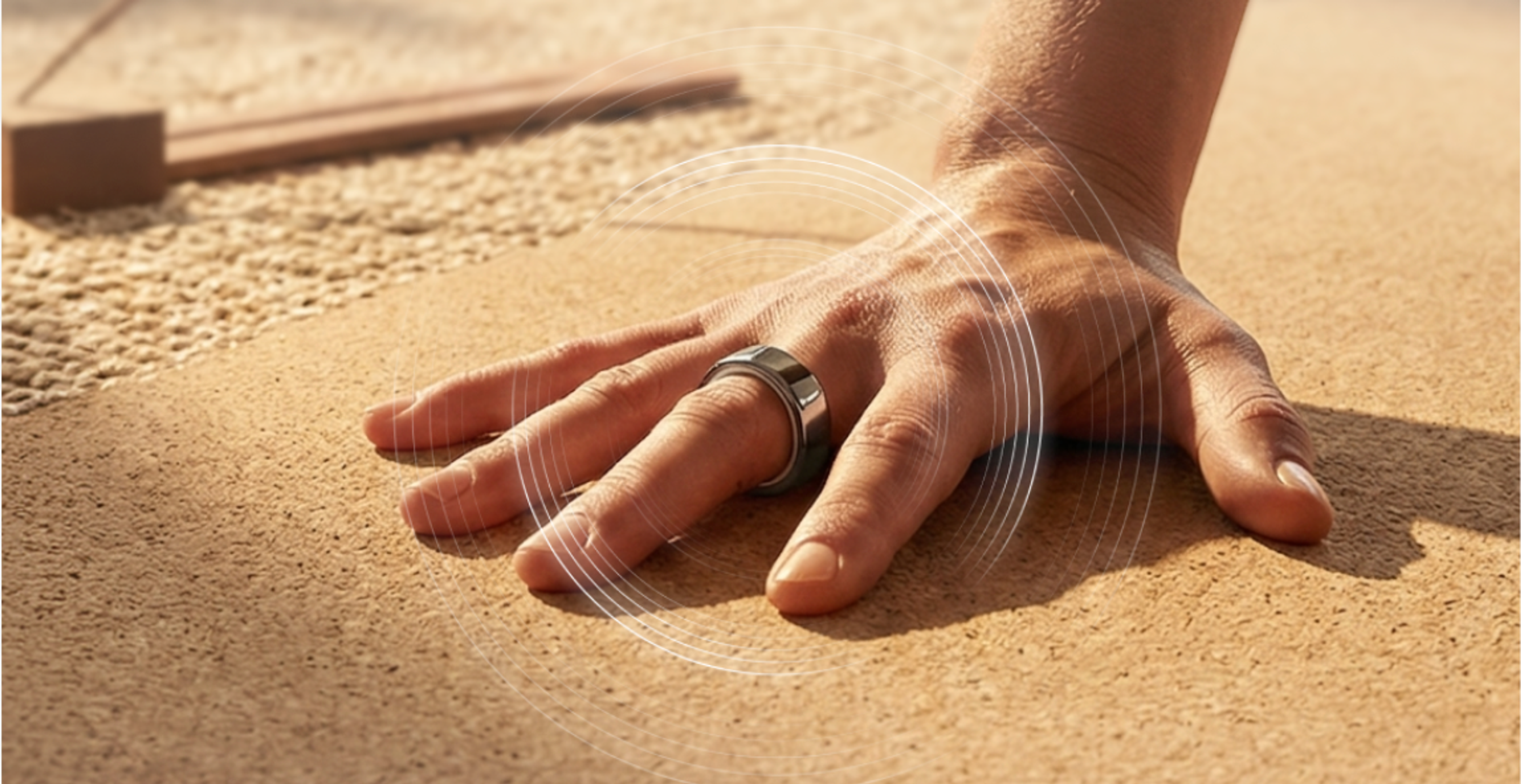 A hand wearing a silver smart ring on the index finger rests on a textured brown surface, with white signal waves radiating from the ring.