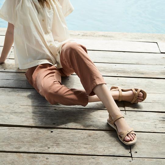 A person wearing a light shirt, rust pants, and tan sandals sits on a wooden dock next to water.