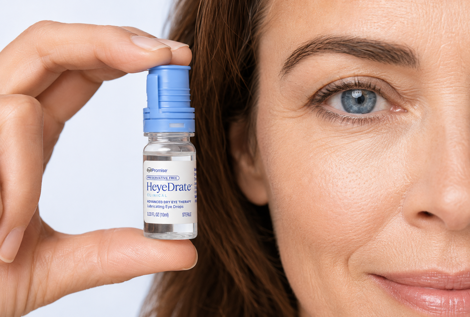 A woman holds a bottle of HeyeDrate Clinical Lubricating Eye Drops next to her blue eye.