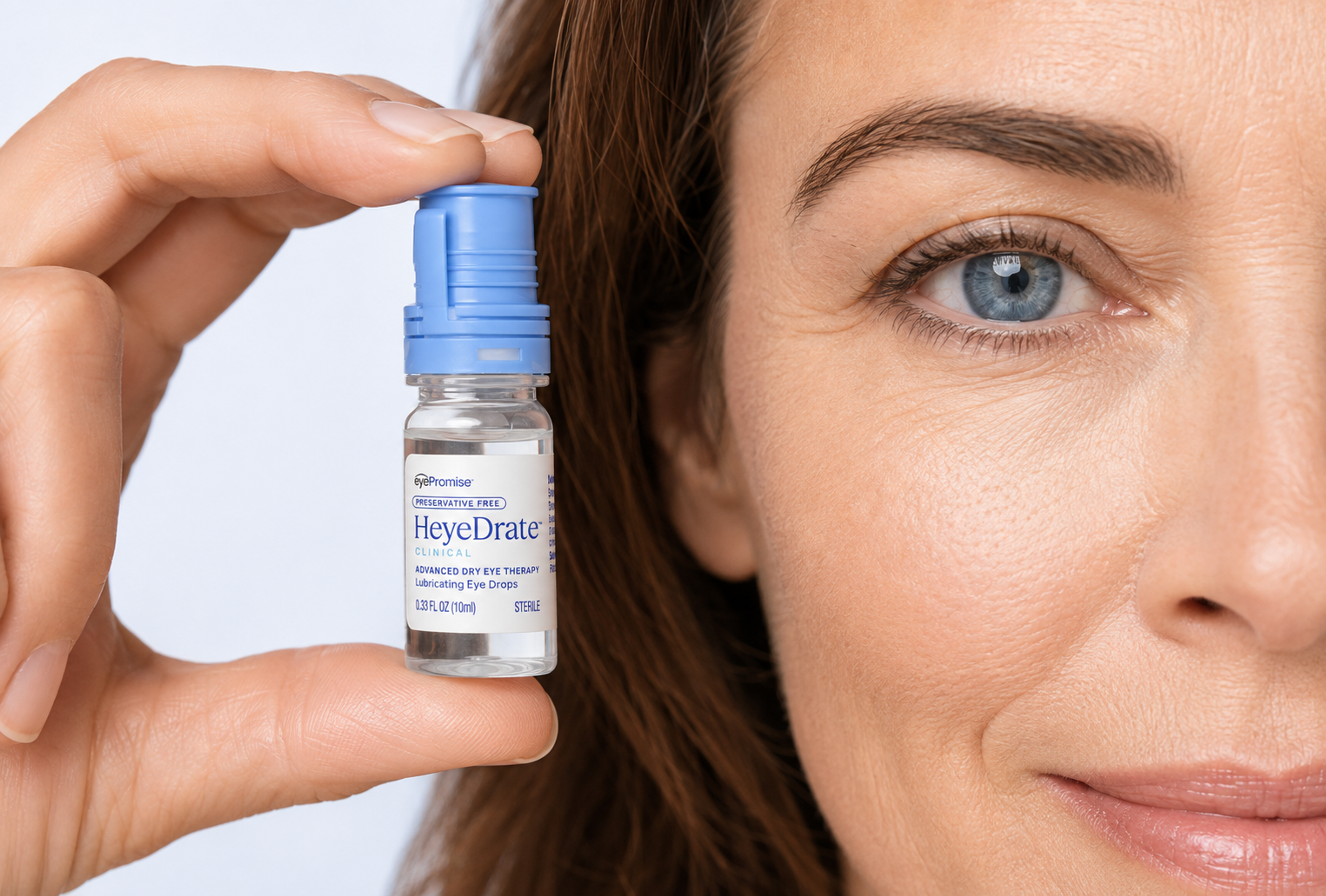 A woman holds a bottle of HeyeDrate Clinical Lubricating Eye Drops next to her blue eye.