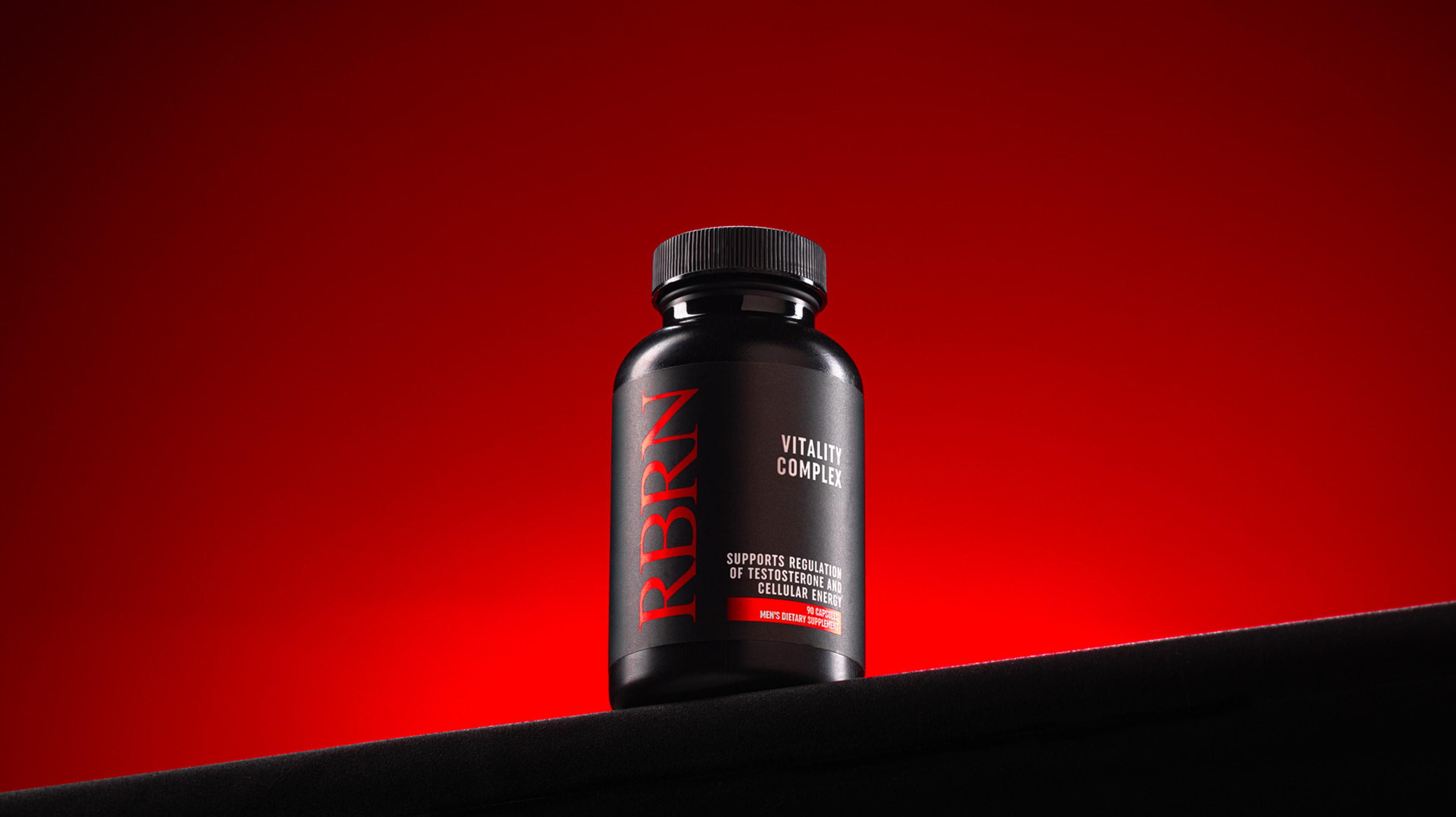A black bottle of RBRN Vitality Complex supplements on a dark surface against a red background.