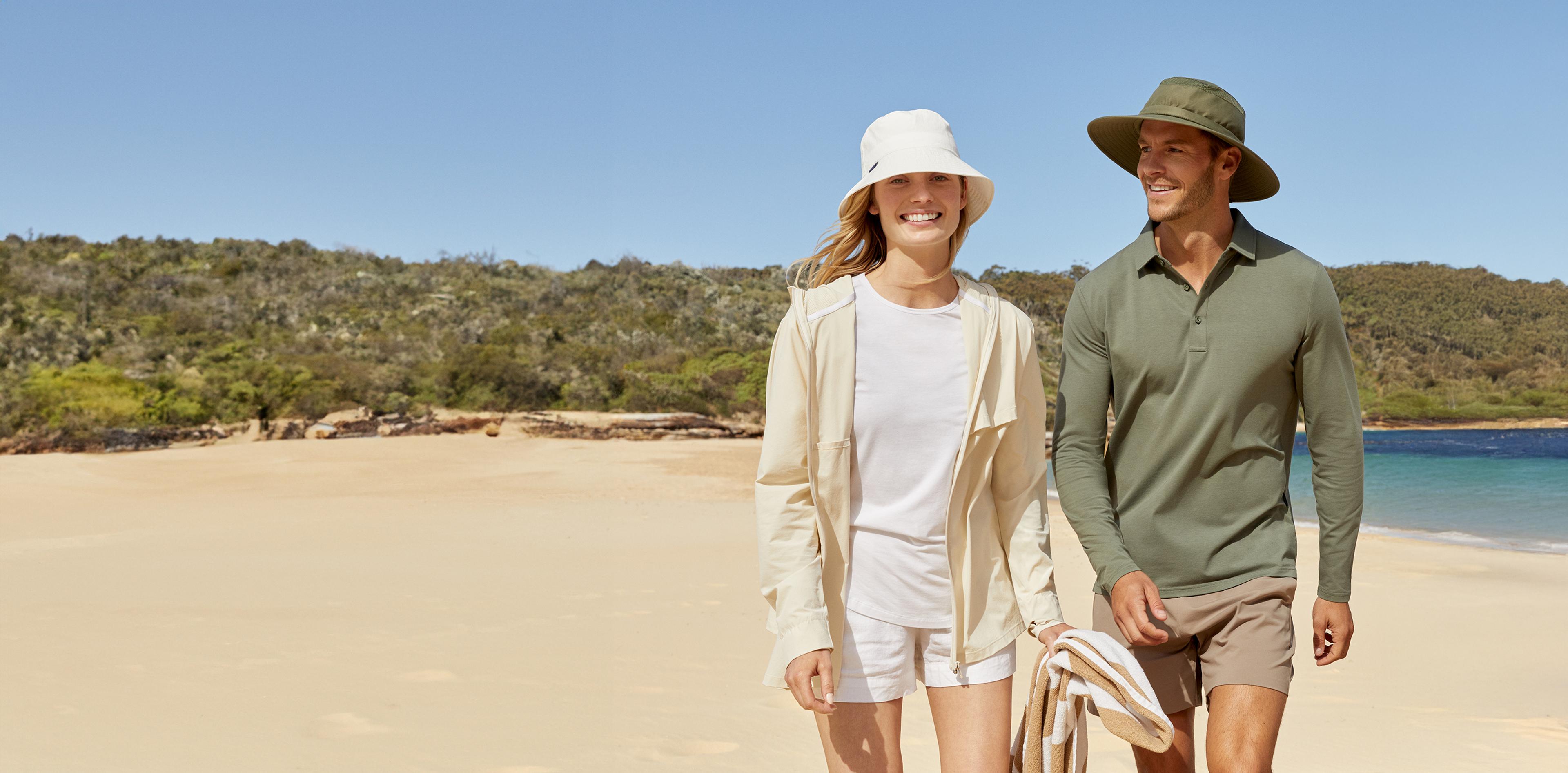 A smiling man and woman wearing sun hats and casual sun-protective clothing walk on a sandy beach.