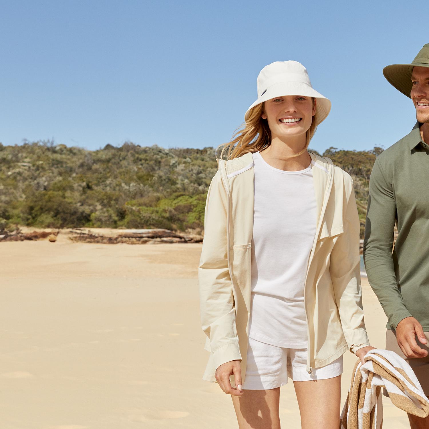 A smiling man and woman wearing sun hats and casual sun-protective clothing walk on a sandy beach.