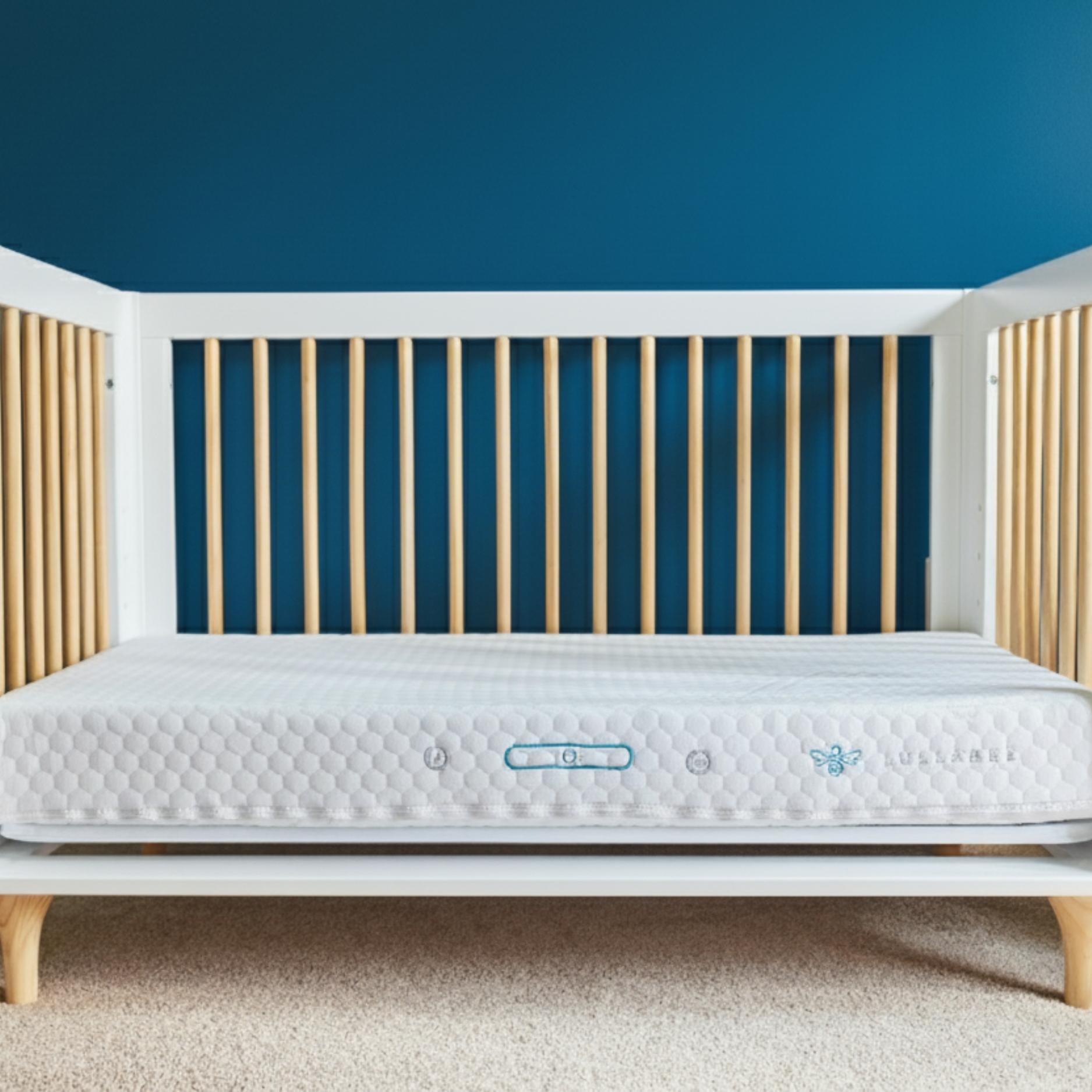 A white crib with natural wood slats and a white mattress with a hexagonal pattern against a blue wall.