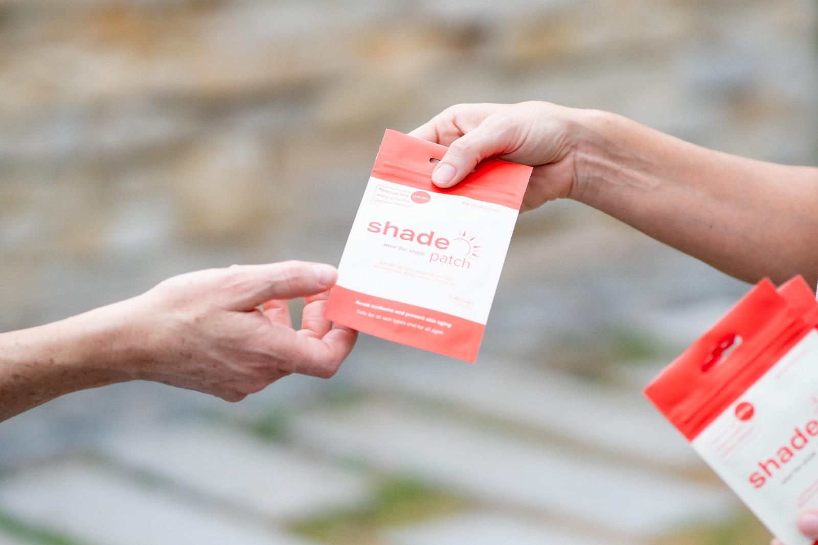 A hand offers a red and white "Shade patch" package to another hand.