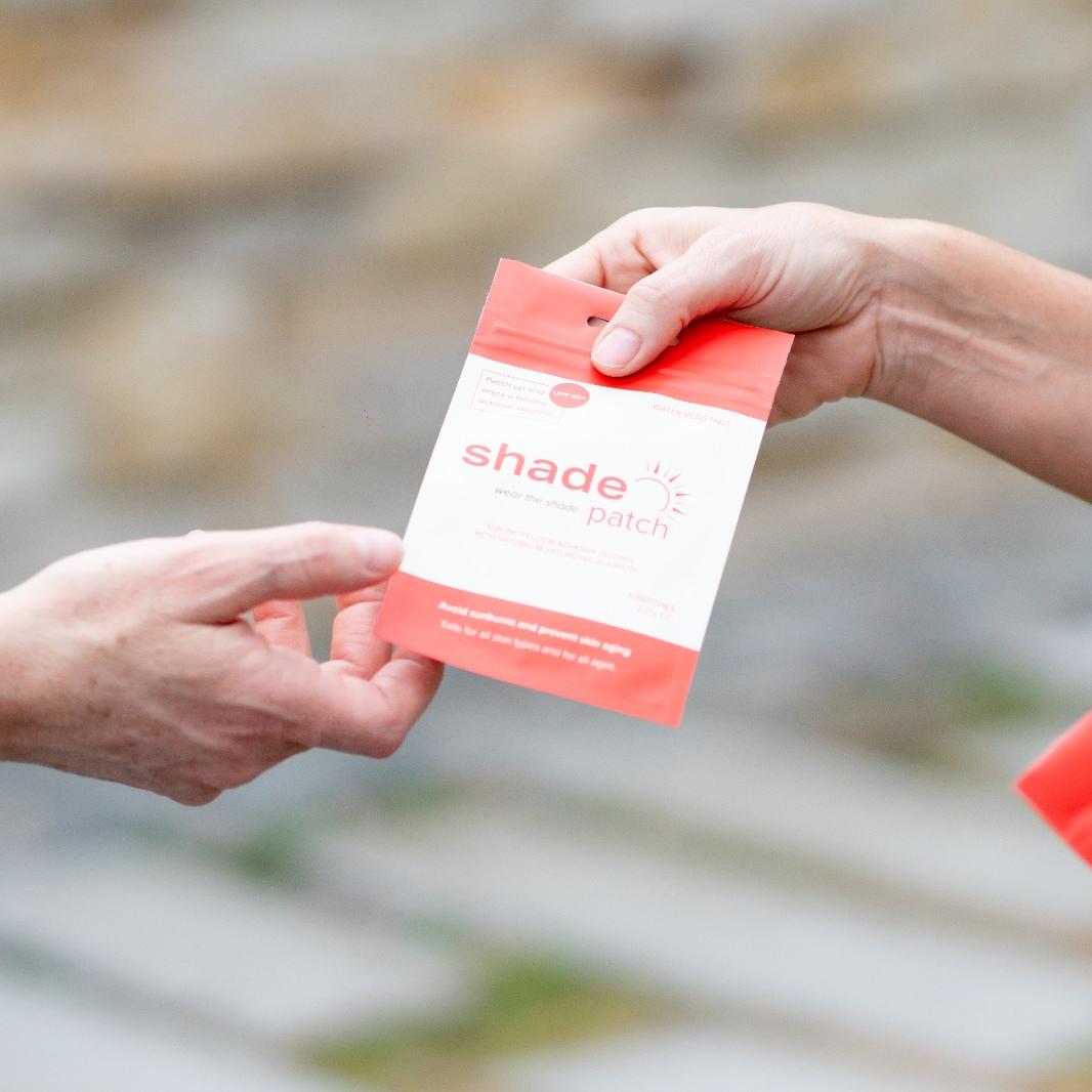 A hand offers a red and white "Shade patch" package to another hand.