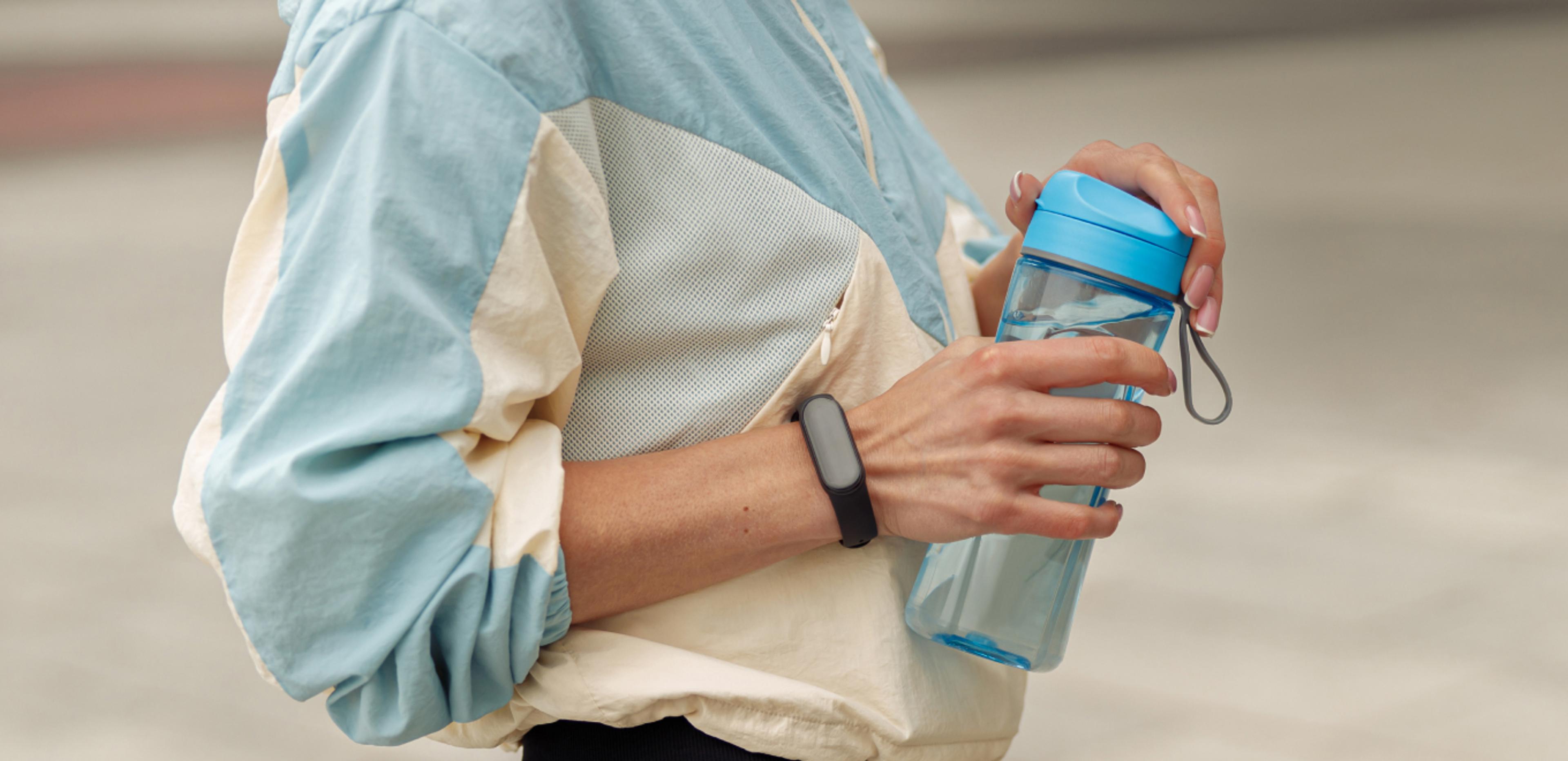 Person wearing athletic clothing and a fitness tracker while holding a reusable water bottle outdoors.