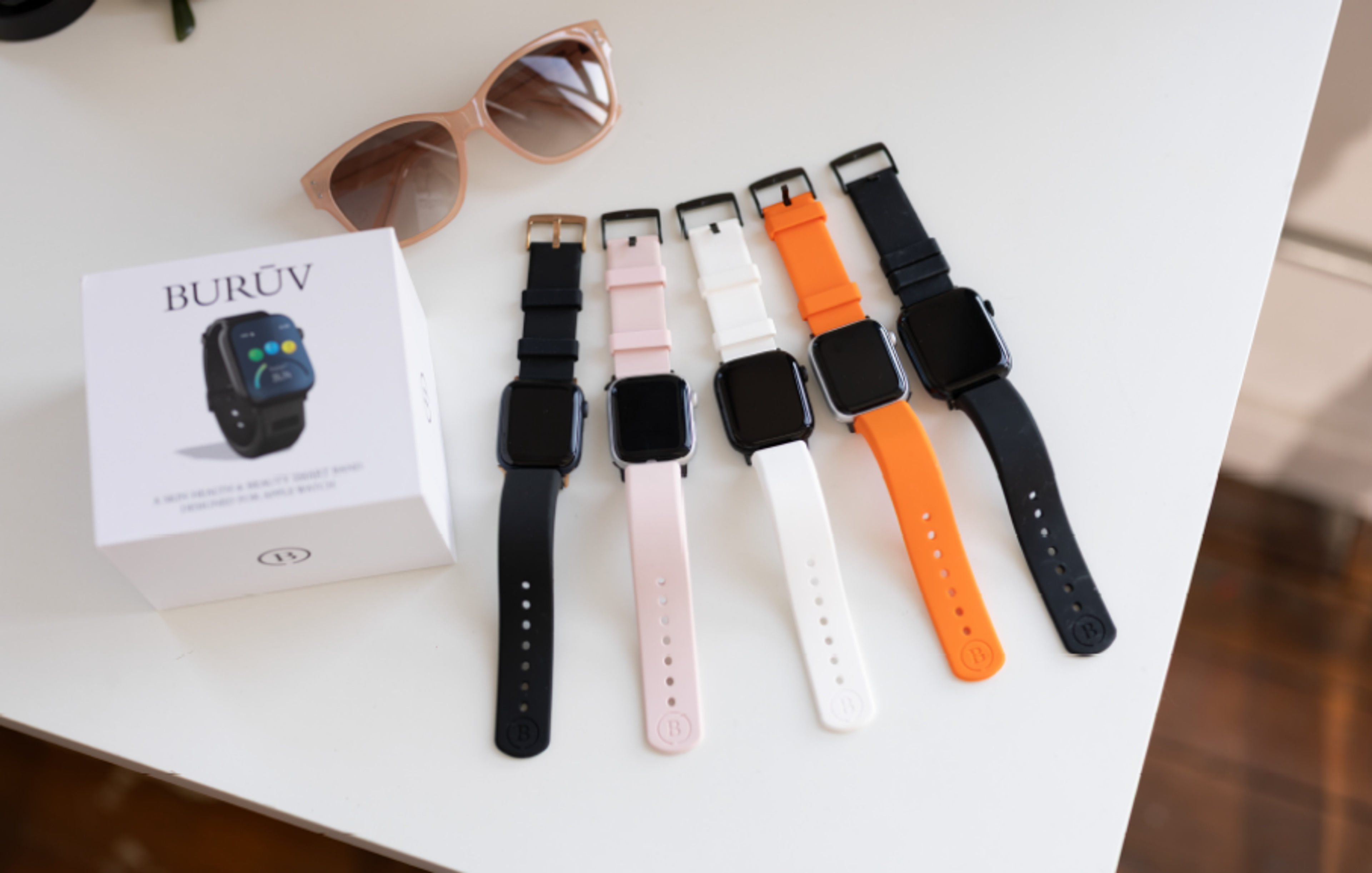 BURŪV smart watch box, sunglasses, and five smart watches with black, pink, white, and orange bands.