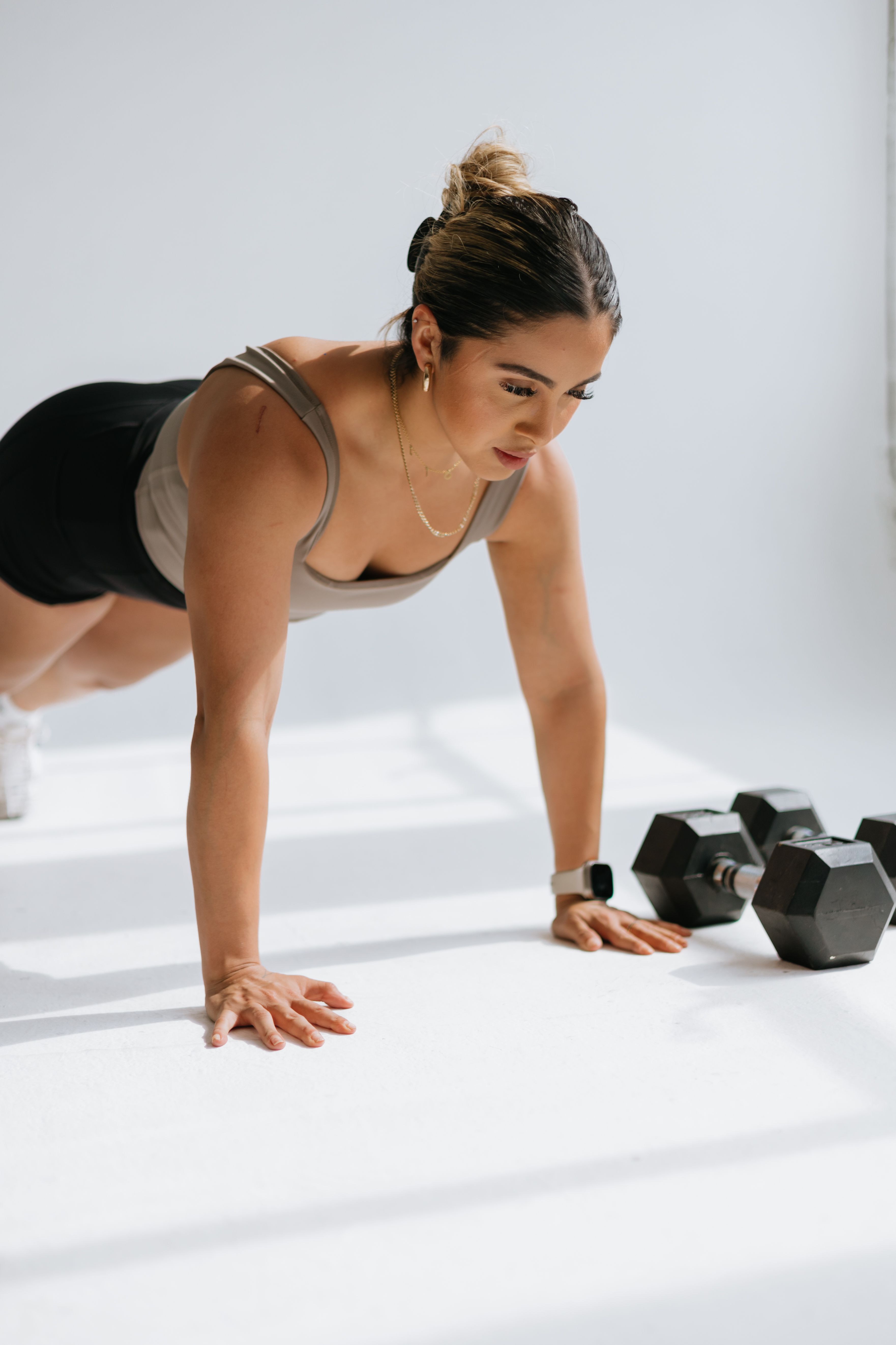 A woman in a plank position on a white floor, with two dumbbells beside her.
