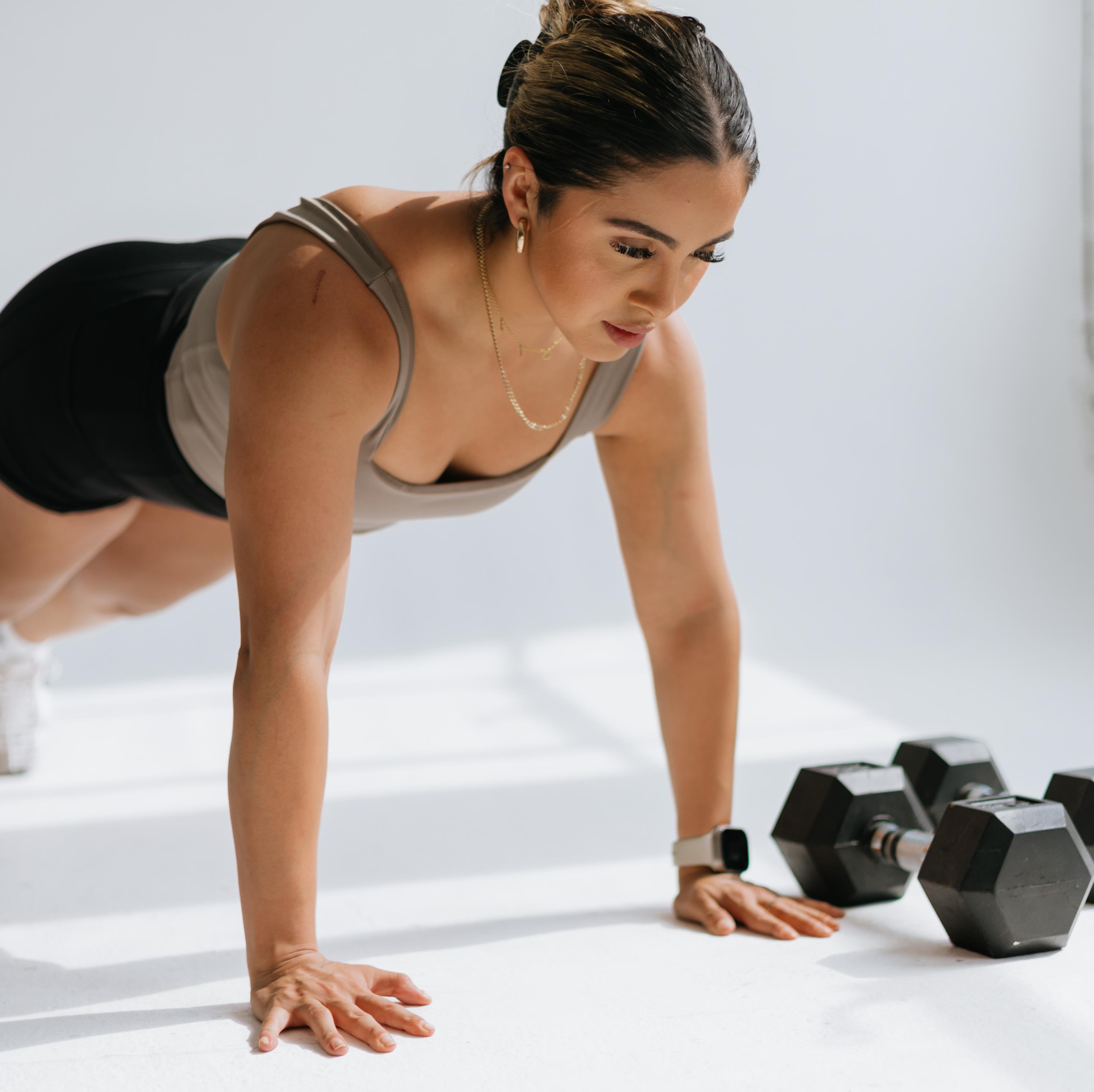 A woman in a plank position on a white floor, with two dumbbells beside her.