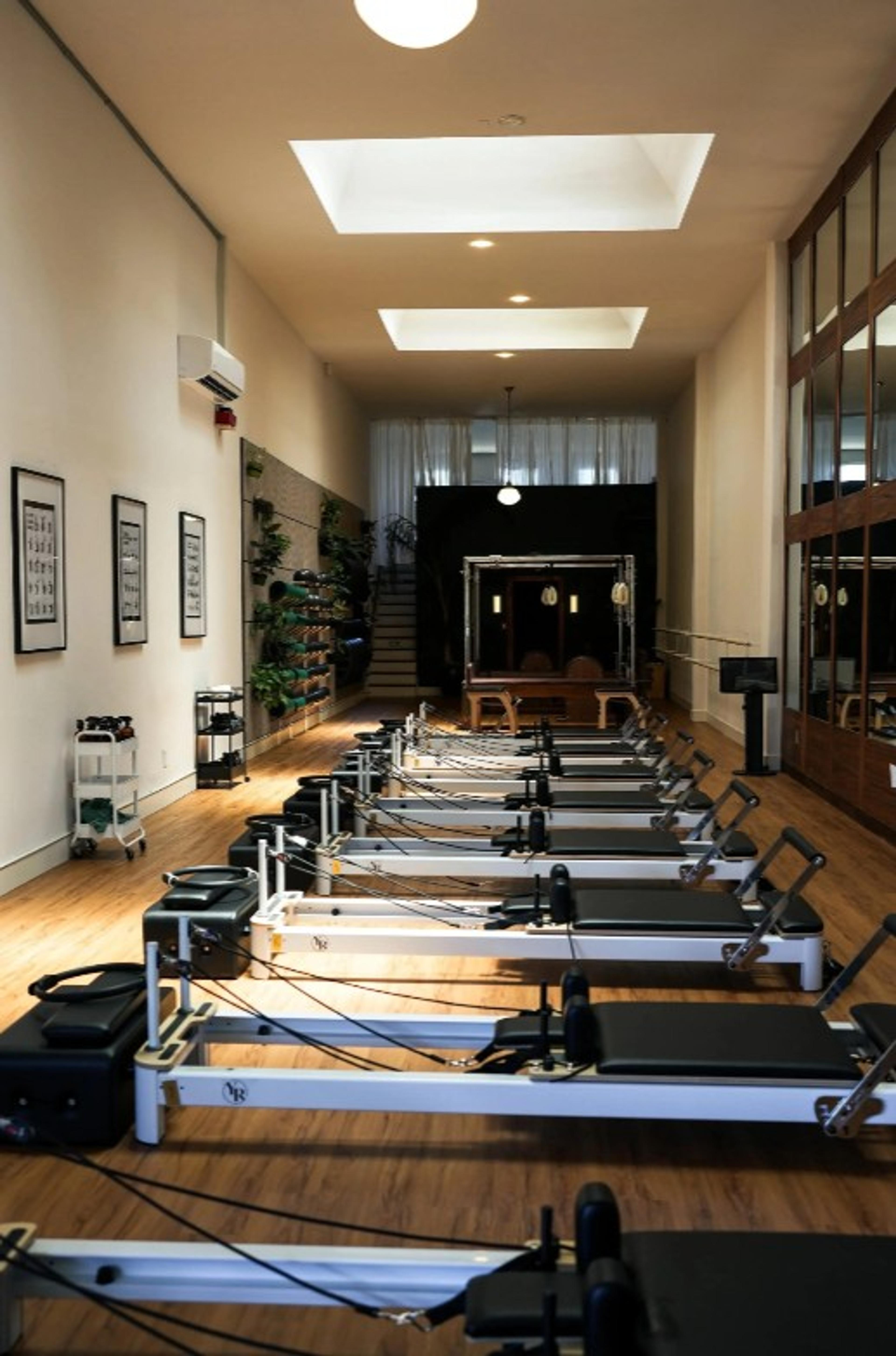 Multiple pilates reformers line a long studio with wooden floors, leading to a darker back area.