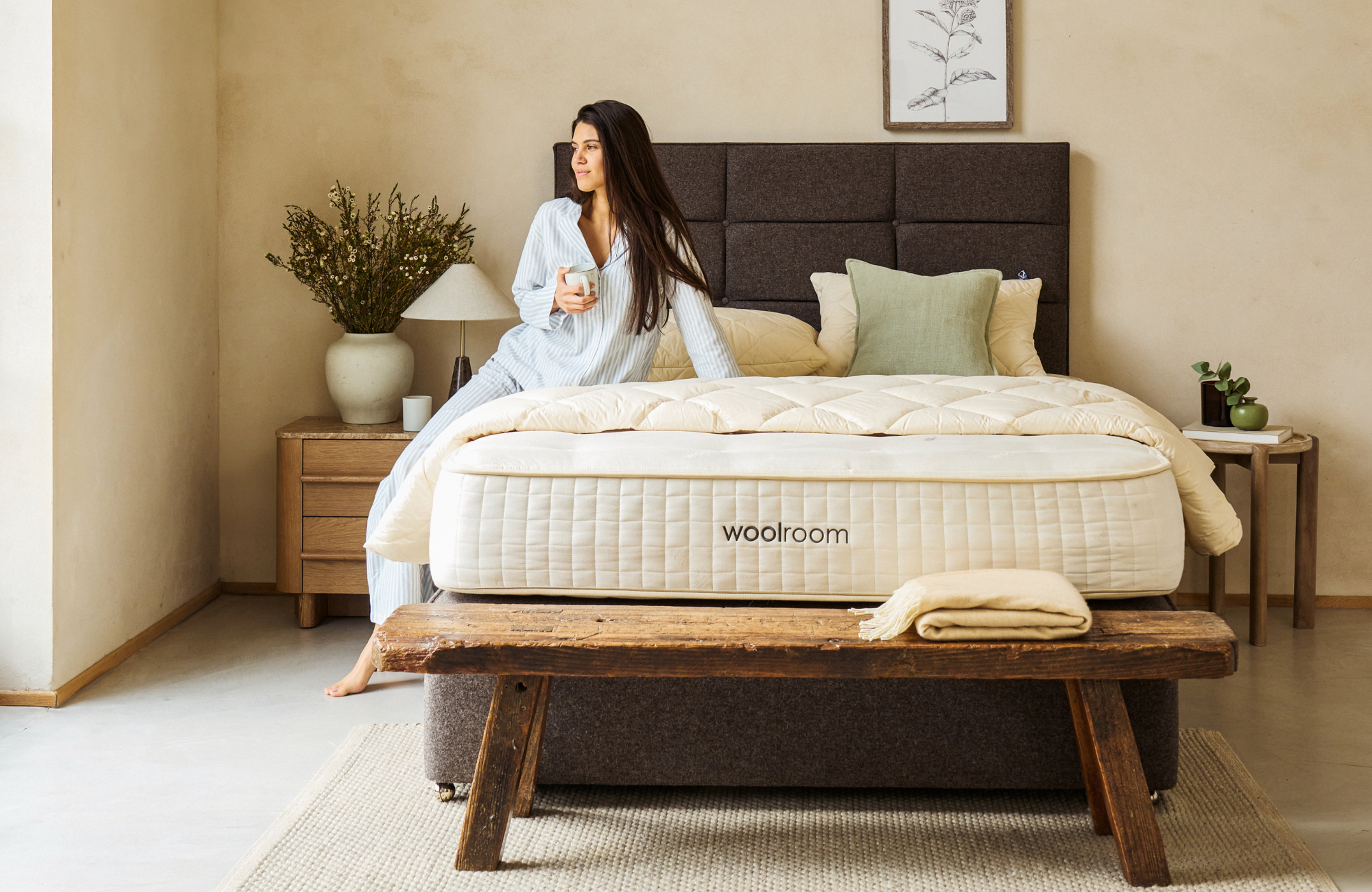 A woman in pajamas sits on a Woolroom mattress, holding a mug and looking out a window in a cozy bedroom.