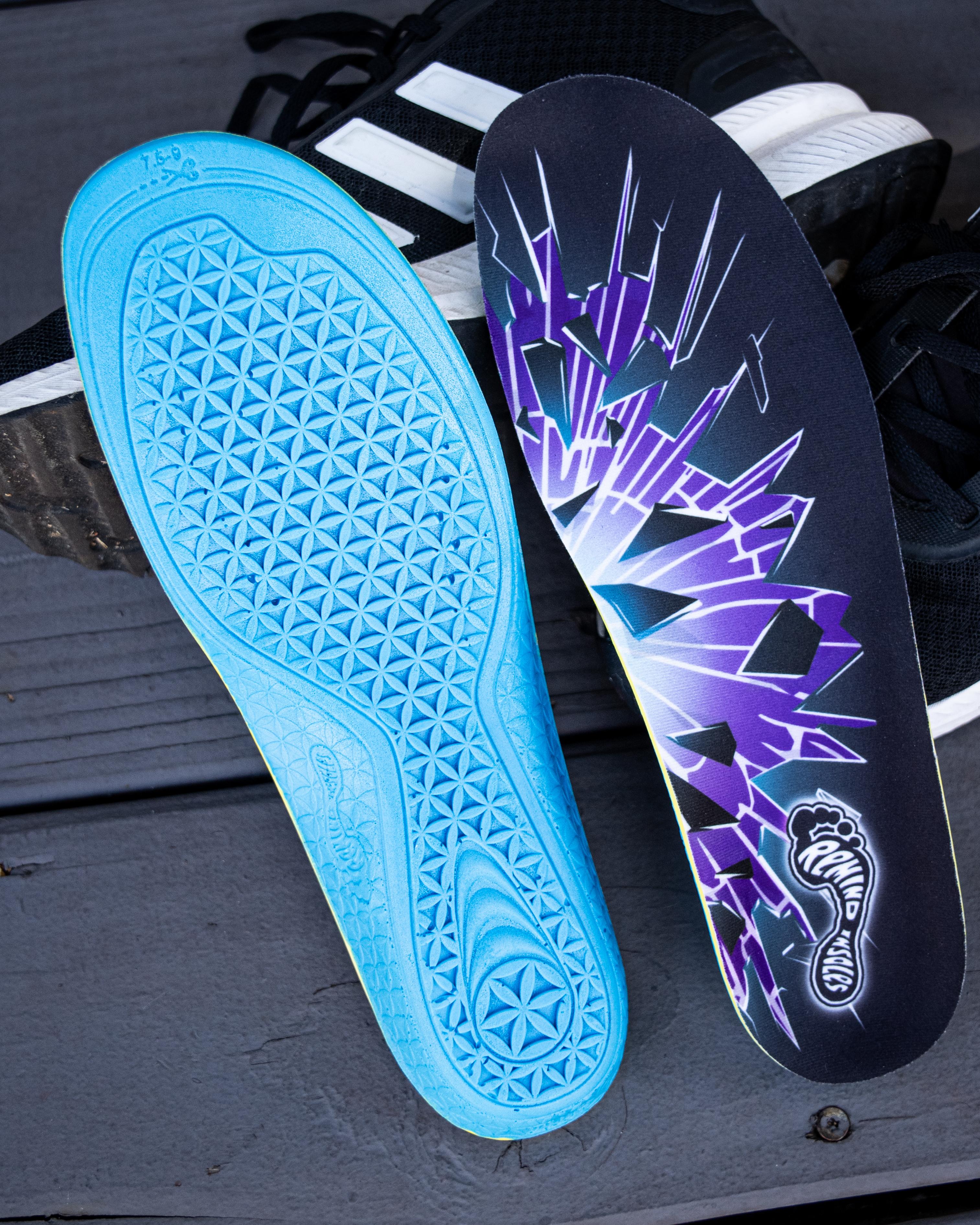 A blue insole with a textured geometric pattern on its base, next to a black insole featuring a purple shattered design and 'Remind Insoles' logo, on a dark wooden surface.