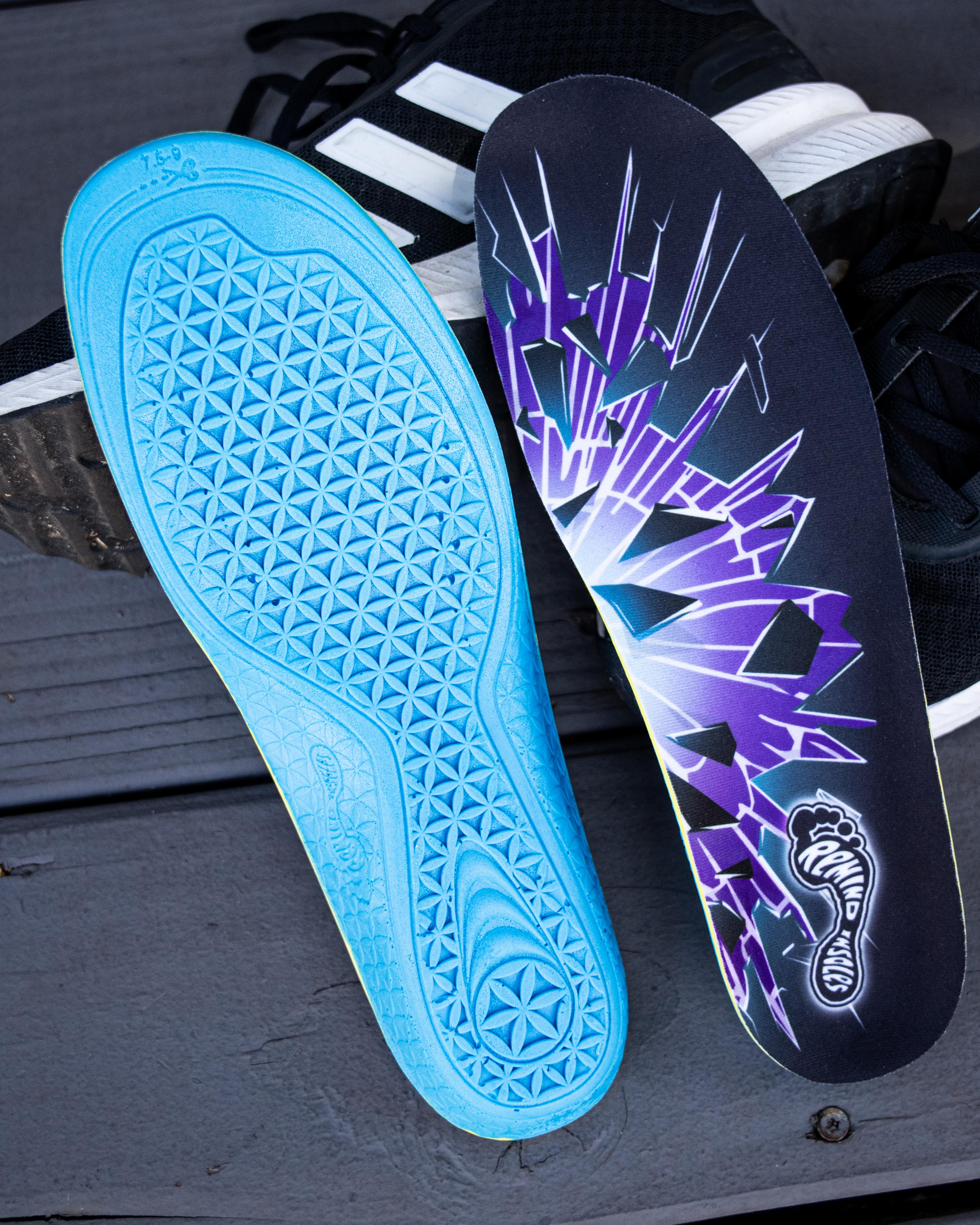 A blue insole with a textured geometric pattern on its base, next to a black insole featuring a purple shattered design and 'Remind Insoles' logo, on a dark wooden surface.