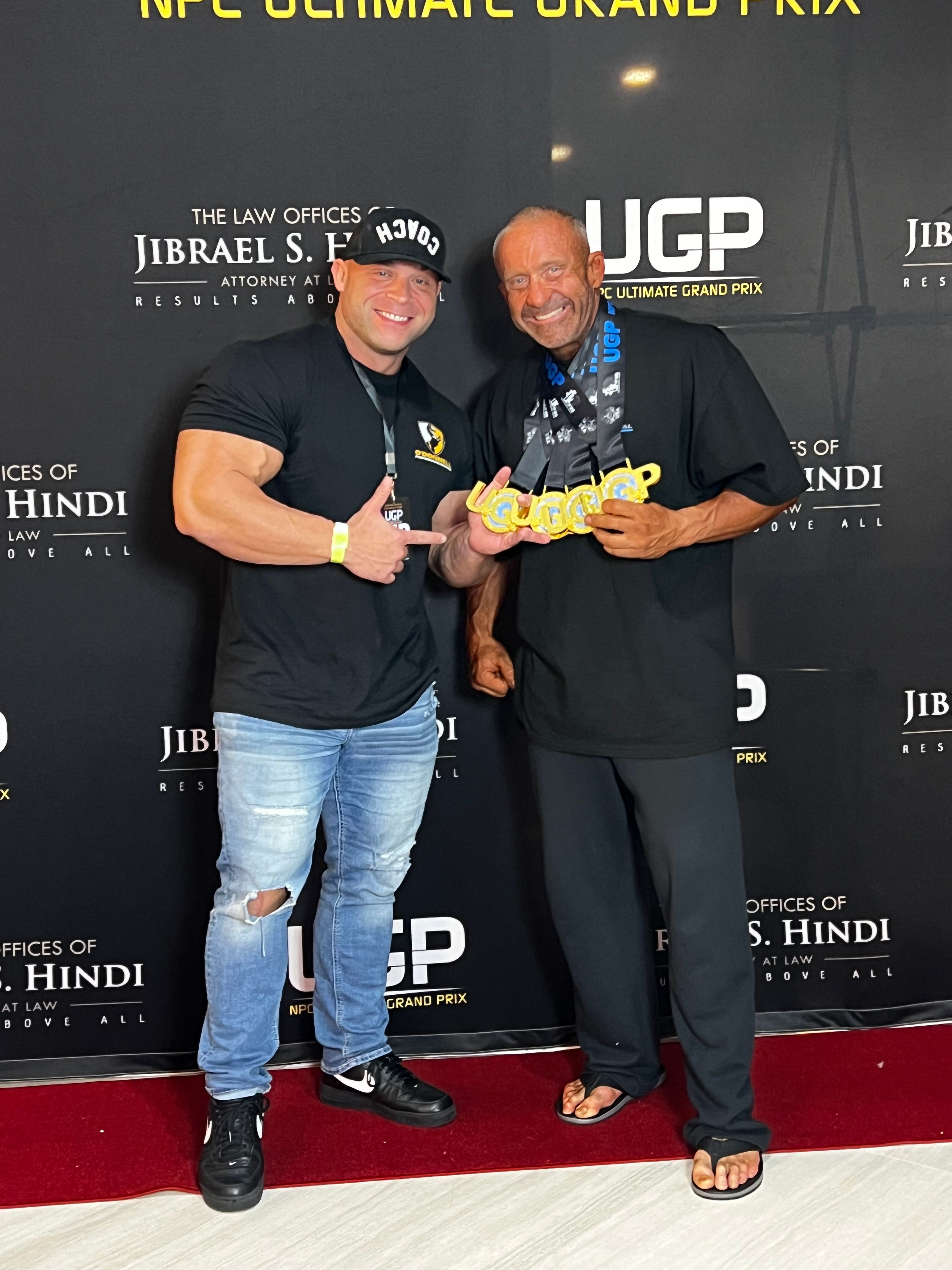 Two smiling men, one a coach giving a thumbs-up and the other an older bodybuilder holding multiple gold medals, pose in front of a sponsor backdrop.