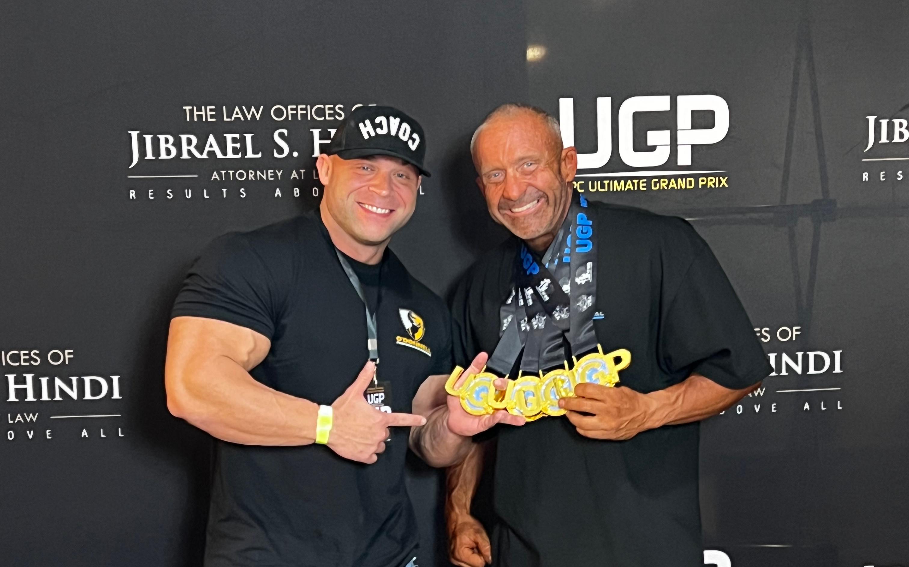 Two smiling men, one a coach giving a thumbs-up and the other an older bodybuilder holding multiple gold medals, pose in front of a sponsor backdrop.