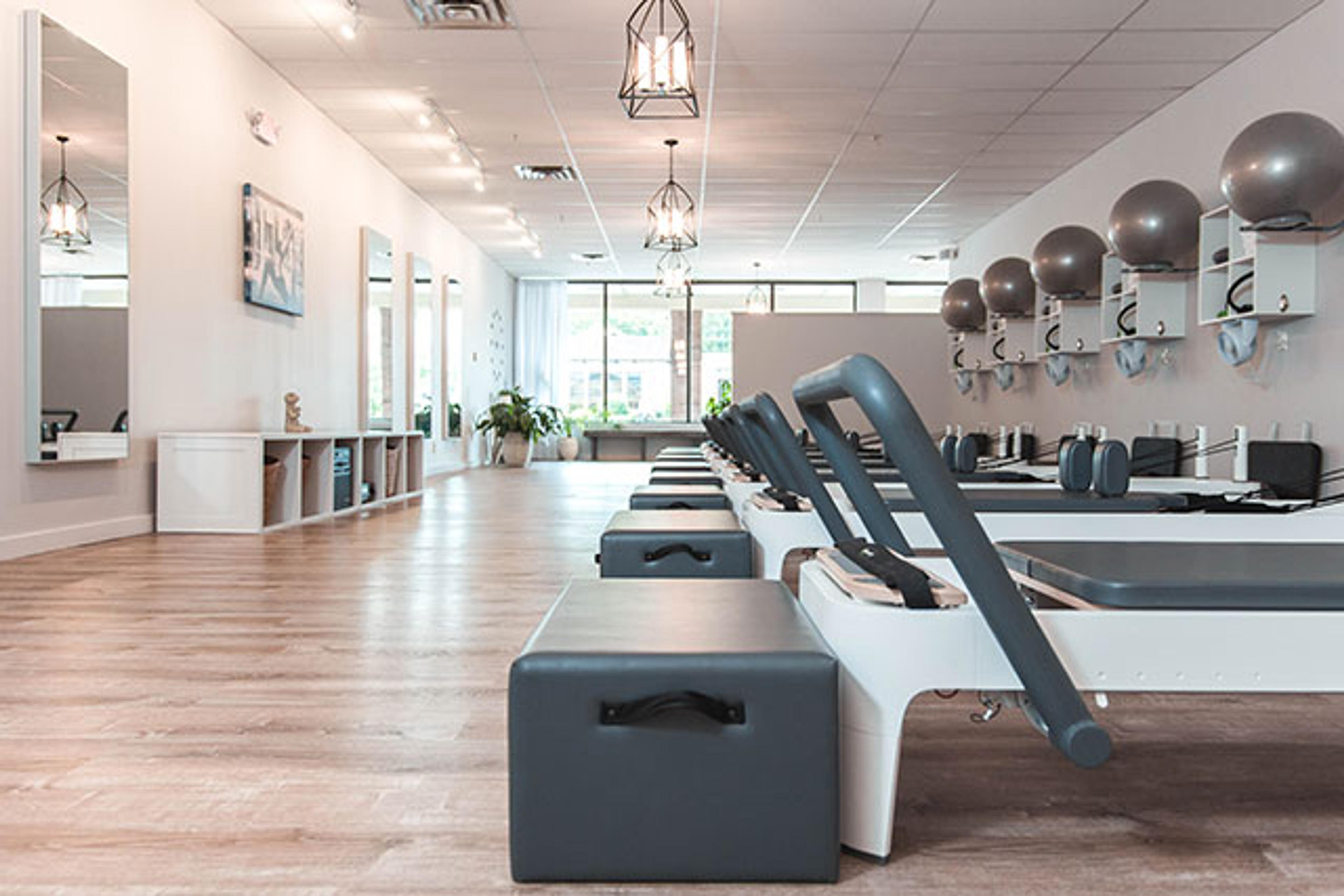 A bright, clean pilates studio with a row of reformers, exercise balls on shelves, and light wood flooring.