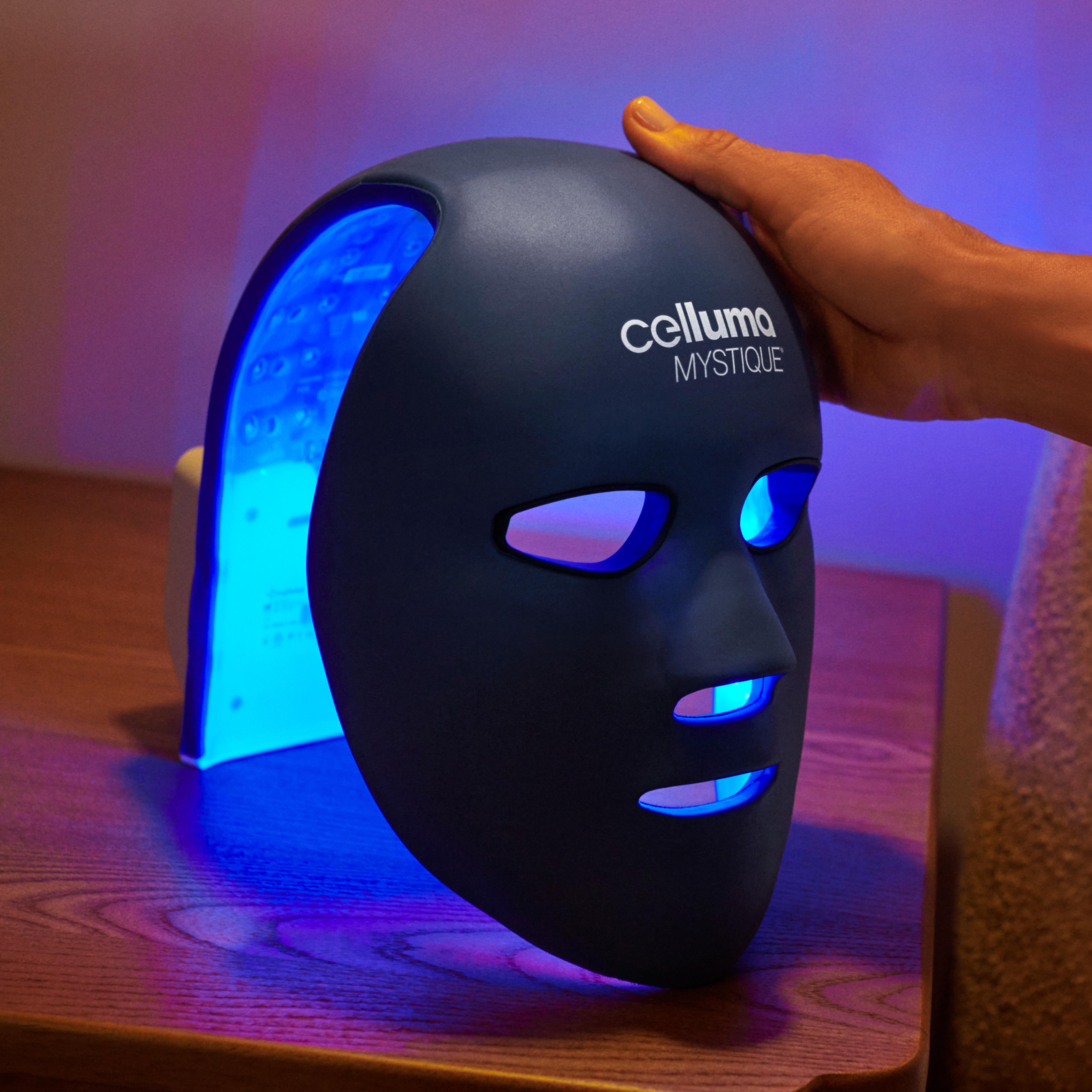 A dark blue Celluma Mystique LED light therapy mask glowing with blue light, a hand resting on its top.